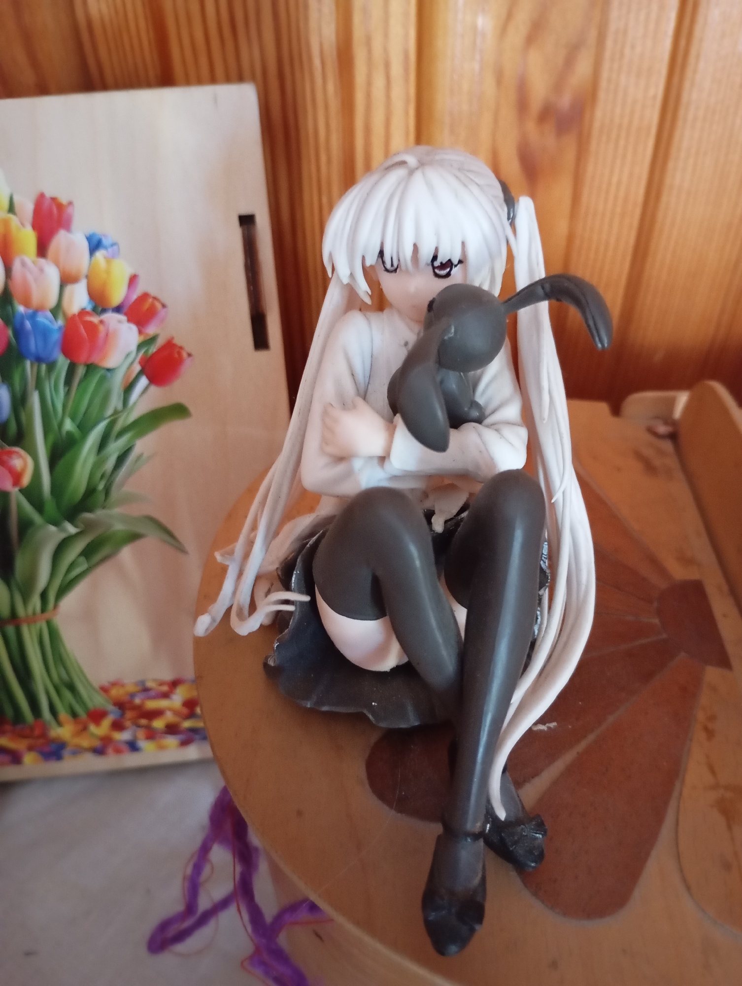 This is a close-up photograph of a figurine of a young girl with long white hair, wearing dark stockings and a light-colored top. She is holding a dark-colored stuffed bunny. The figurine is seated on a wooden surface with a patterned inlay. In the background, to the left of the figurine, is a bouquet of colorful tulips. The overall background consists of a wooden wall. This scene does not provide any specific geographical or urban context that would allow for identification of its location in L'nikha, Russia.