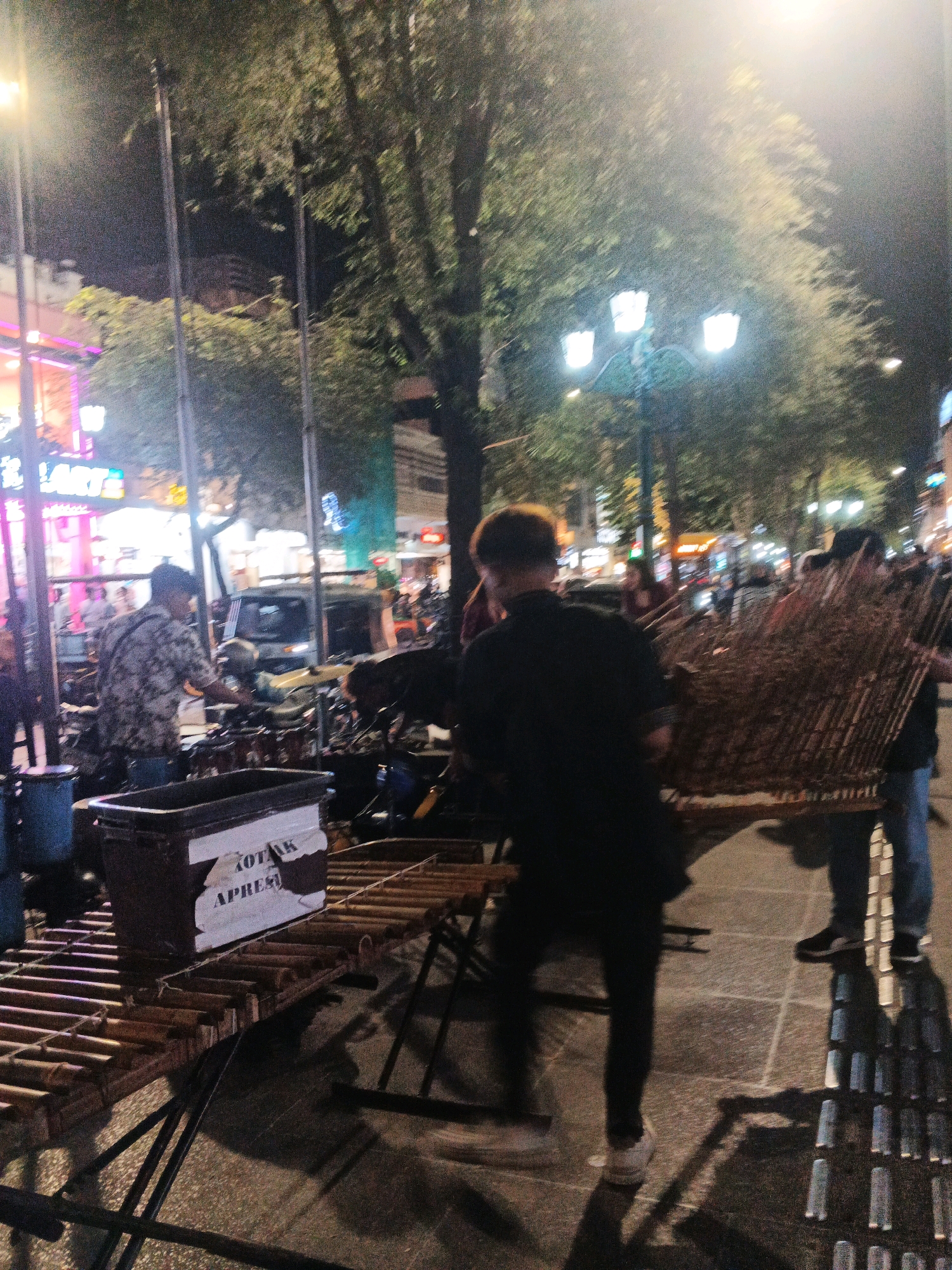 A nighttime street scene in Yogyakarta, Indonesia, shows an outdoor performance or setup. In the foreground, a person in a black shirt and pants is facing away from the camera, holding a large, bundle of what appears to be dried reeds or bamboo, possibly part of a musical instrument or stage decoration. To the left, another person in a patterned shirt is partially visible, appearing to interact with a drum set or percussion instruments laid out on a wooden platform. A black plastic container with white text reading "KOTAK APRES" is placed on the platform. The background reveals a street lined with trees, illuminated by streetlights and neon signs from buildings. The setting suggests a public space, possibly a pedestrian area or side street in a commercial district of the city.