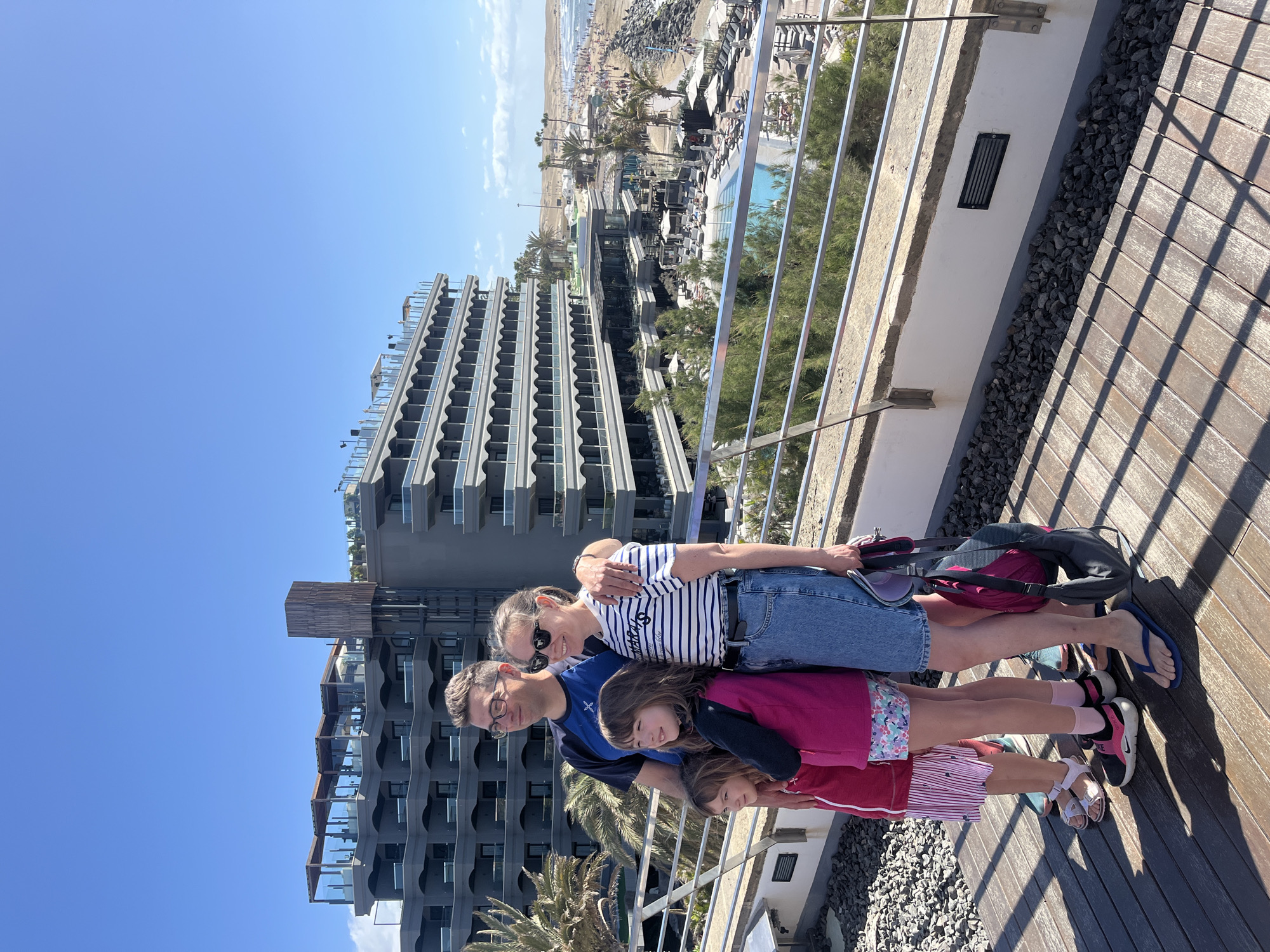 A horizontally-oriented medium shot presents a family of four—two adults and two children—standing shoulder-to-shoulder on a wooden deck, with an urban scene in the background. The adults, presumably parents, stand at the top of the frame, the man on the left with eyeglasses and the woman on the right with sunglasses, her arm around his shoulder. Below them, two girls, presumably their daughters, stand close together. The background showcases a multi-story, modern architectural building with repeating balcony patterns under a clear blue sky. In the distance, a beach and resort area with palm trees, pools, and people can be seen. The deck is lined with a metal railing and small gravel along the side.
