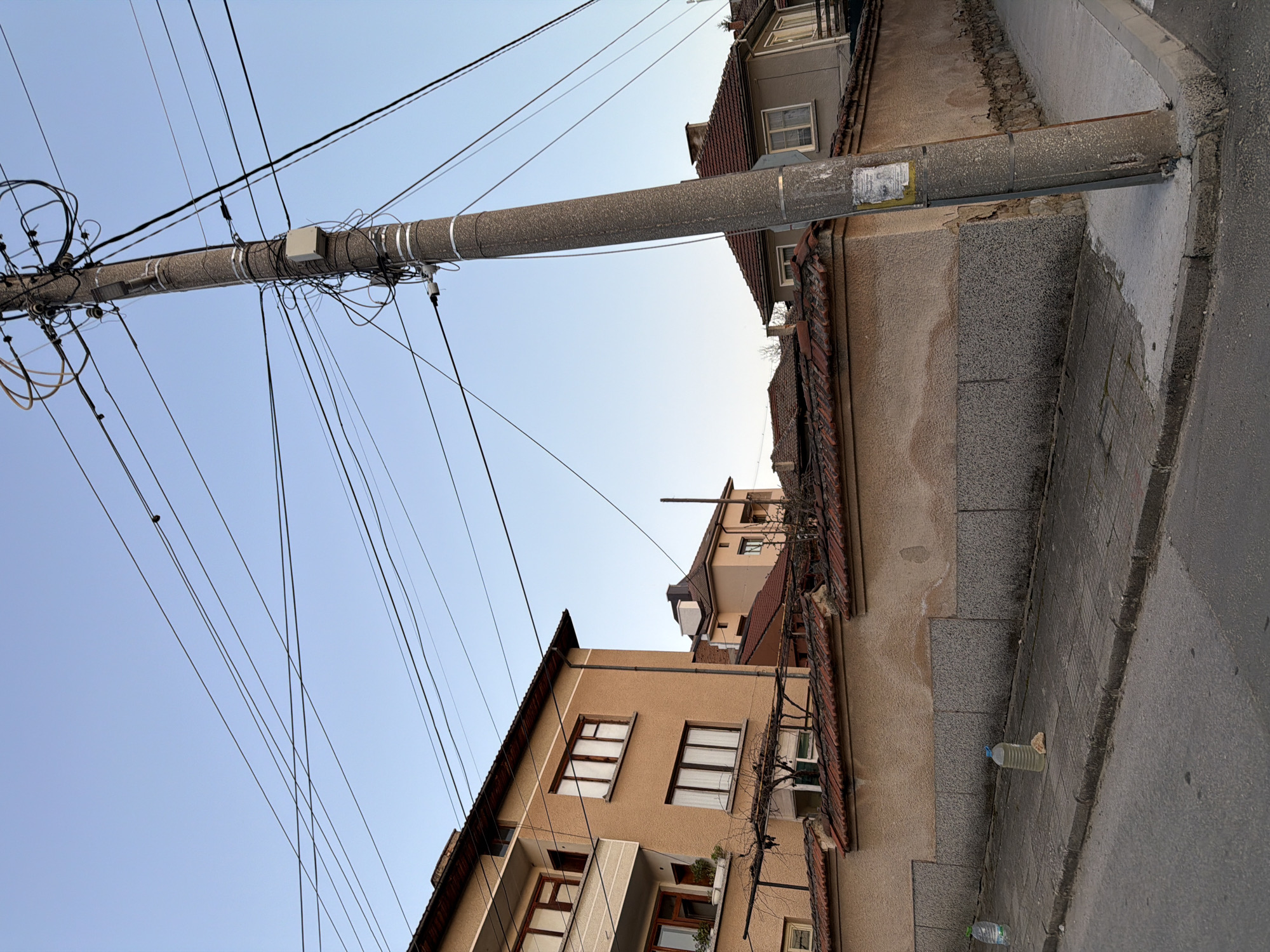 This photograph, taken in Panagyurishte, Bulgaria, shows a street scene with a low-angle perspective. A concrete utility pole extends horizontally across the frame, heavily laden with electrical wires. The pole appears to be mounted to the side of a building, near its roofline. The building is multi-storied with a light brown facade and several windows with white frames. The roof is tiled in reddish-brown. The street is paved with gray cobblestones and asphalt. The sky is clear and pale blue. Several plastic bottles are visible at the edge of the road. The photograph gives a sense of cluttered infrastructure and urban architecture.