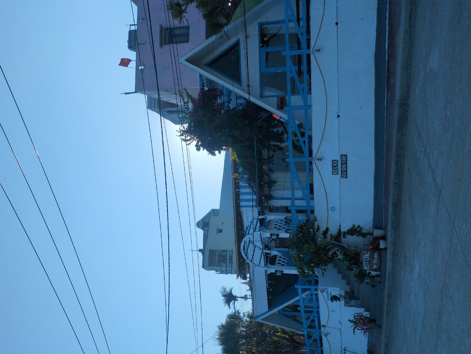 A street-level view in East Quy Nhon Ward, Vietnam, depicts a multi-story building complex, likely a guesthouse or small hotel, under a clear, light blue sky with several overhead power lines. The primary structure features a white facade accented with blue wooden fencing and railings, characteristic of a coastal theme. On the right, a prominent A-frame section contains a triangular window above blue double doors, numbered "B001." To its left, another section of the building includes an arched, white and blue pergola structure, potentially an entrance, with a white wall below featuring the signs "JUST CHILL by the sea" on a wooden plank, and "RELAX SEA-SUN-STAY" on a dark plaque. Steps lead up to an entrance on the left, marked "LA BEACH HOUSE 2," flanked by potted plants and a small, illuminated tree. Bougainvillea vines with purple flowers grow over parts of the building. In the background, a light pink building with a dark window is visible, topped with a red Vietnamese flag featuring a yellow star and a water tank. Further back and to the left, a light-colored multi-story building with balconies and palm trees is present. The foreground consists of a gray, paved street or sidewalk. No individuals or active interactions are visible in the scene.
