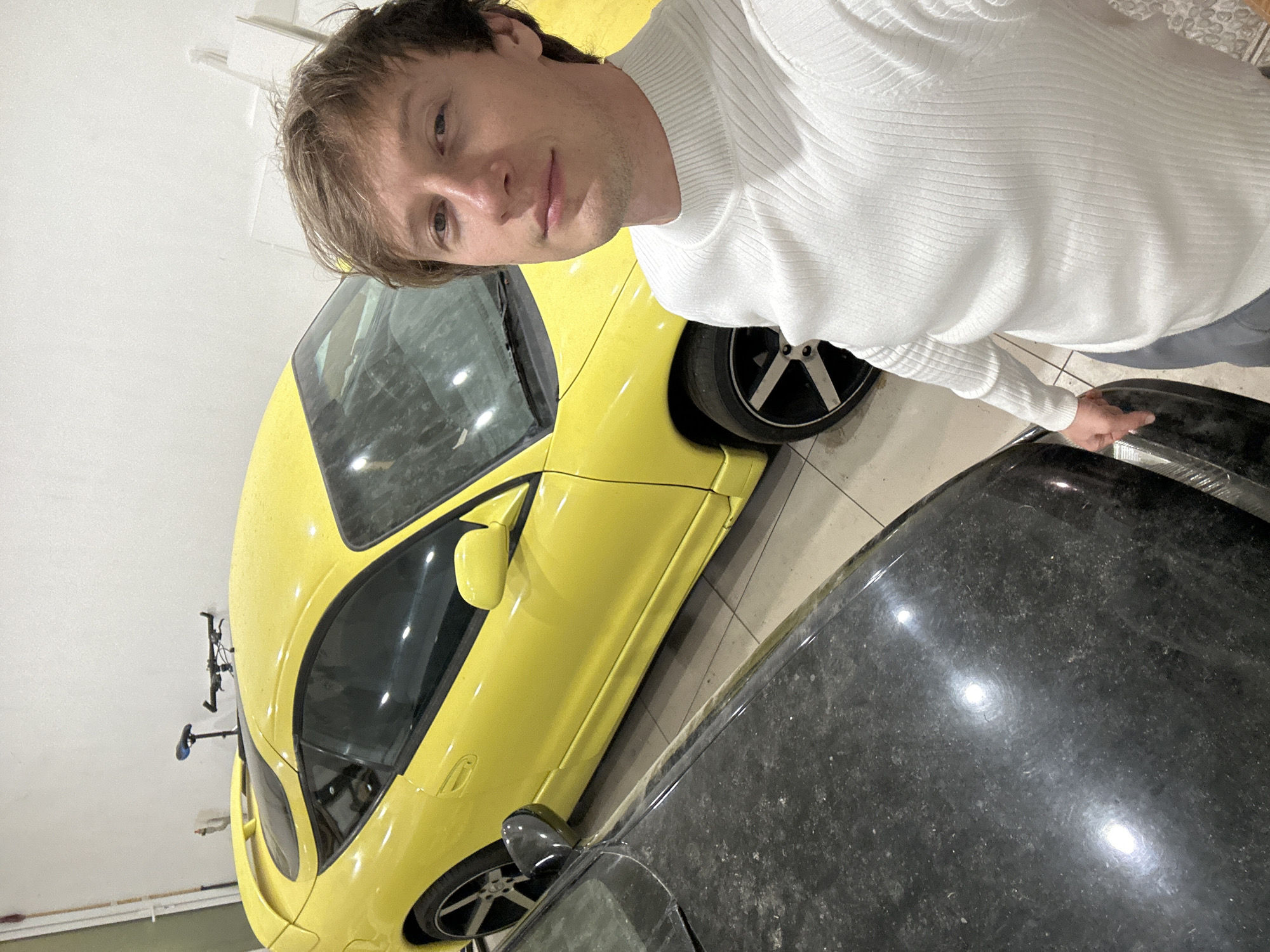 A man wearing a white turtleneck sweater is standing next to a yellow sports car and a dark-colored car in what appears to be a garage. A bicycle is mounted on the wall behind the yellow car. The floor is tiled. There is no visual information to support a location in Kursk, Russia.
