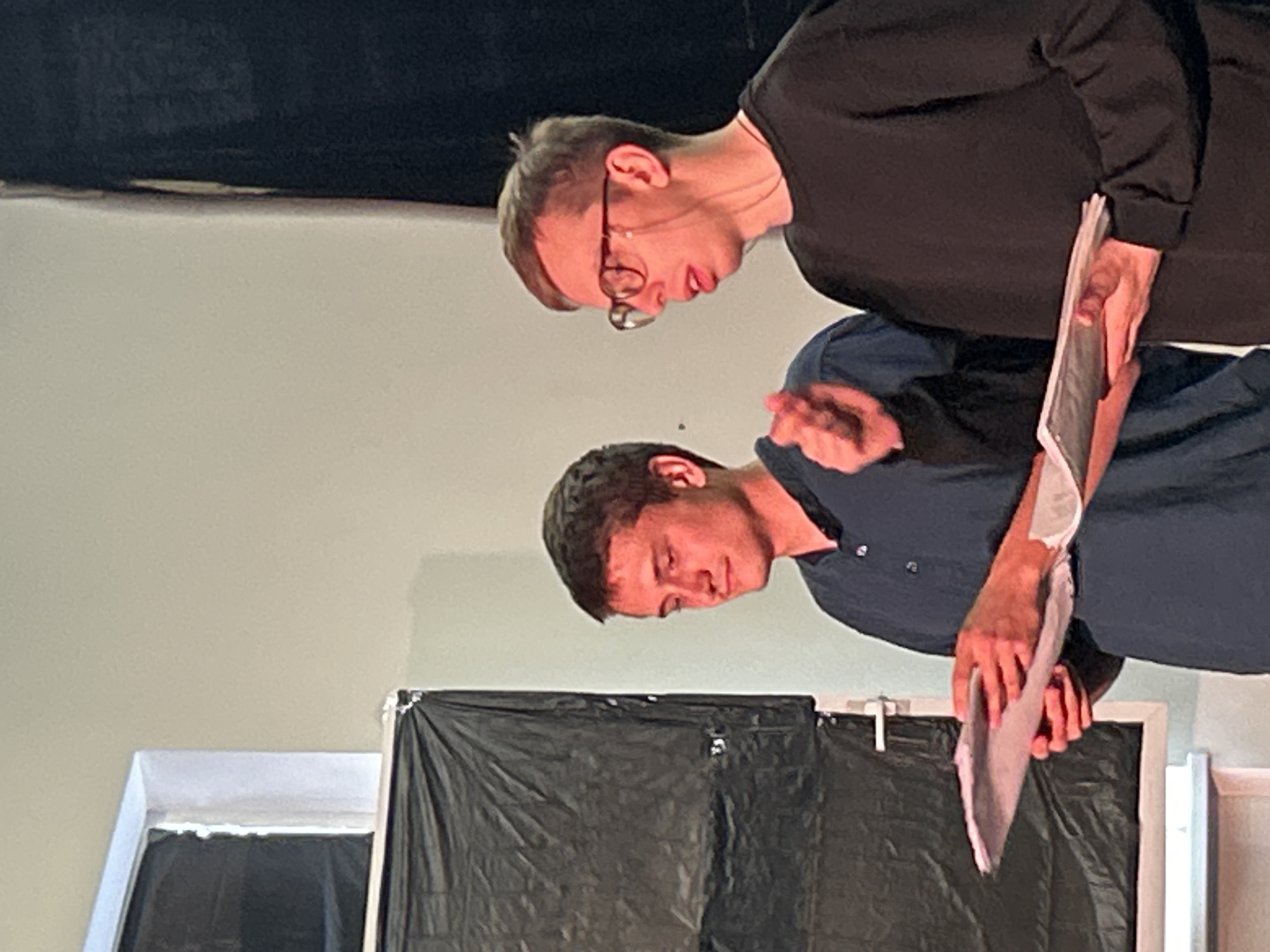 Two young men are standing in front of a black backdrop. The man on the left is wearing a dark blue collared shirt and is holding a stack of papers. He has short brown hair and is looking down at the papers. The man on the right is wearing a black long-sleeved shirt and glasses. He has short brown hair and is looking off to the side. He appears to be speaking and gesturing with his right hand. There is a white window frame to the left of the men, and a white curtain or sheet is hanging from it. The overall scene appears to be indoors, possibly a stage or a rehearsal space.