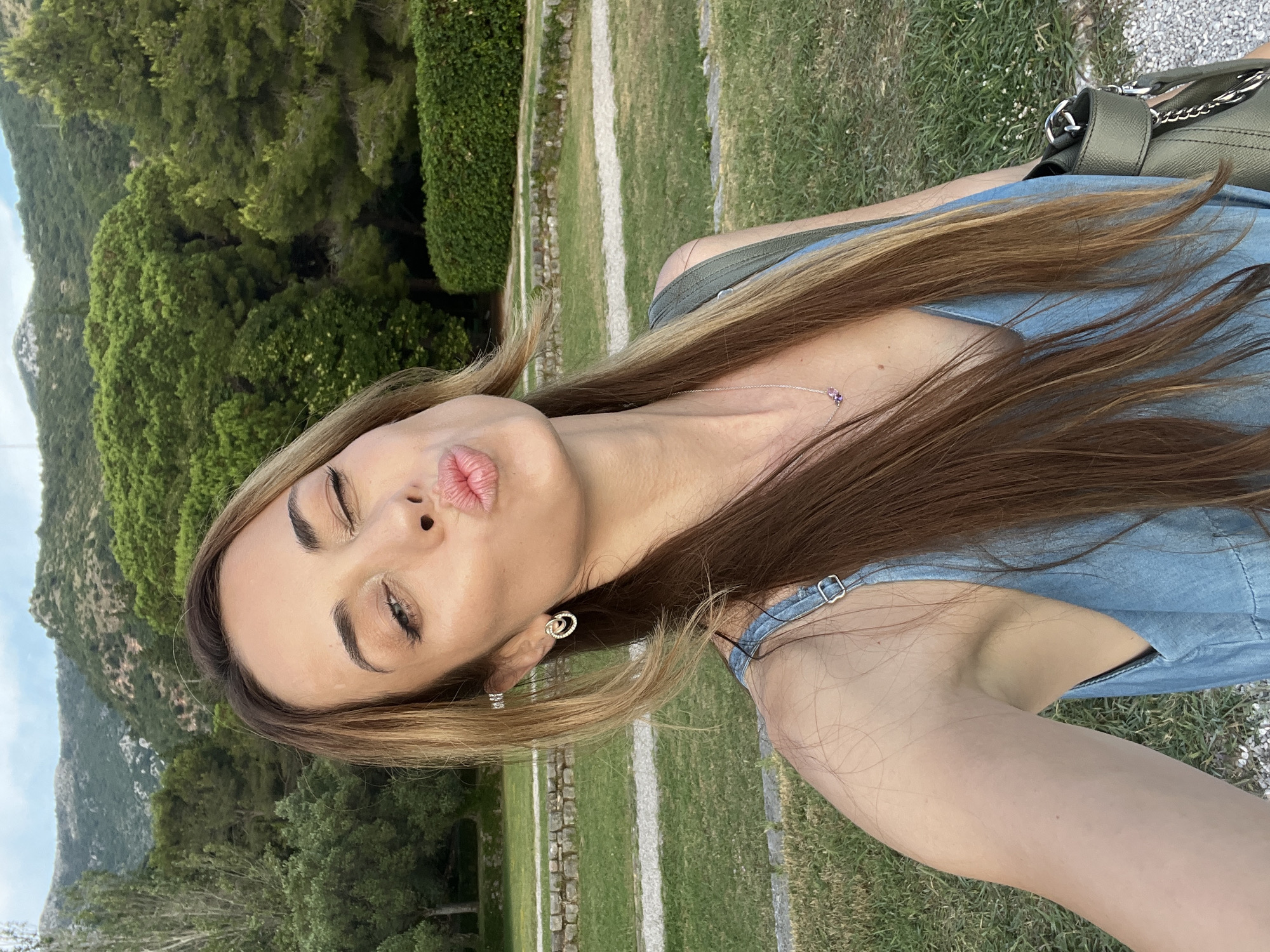 The image is a selfie of a woman with long brown hair, wearing a blue dress, and a green purse. She is making a kissy face and winking. The background is a lush green landscape with trees and hills, suggesting an outdoor location. The environment appears to be a park or garden, with manicured grass and stone pathways. The woman is positioned in the foreground, with the natural scenery behind her. The lighting suggests it is late afternoon or early evening. The location is identified as Sveti Stefan, Montenegro.