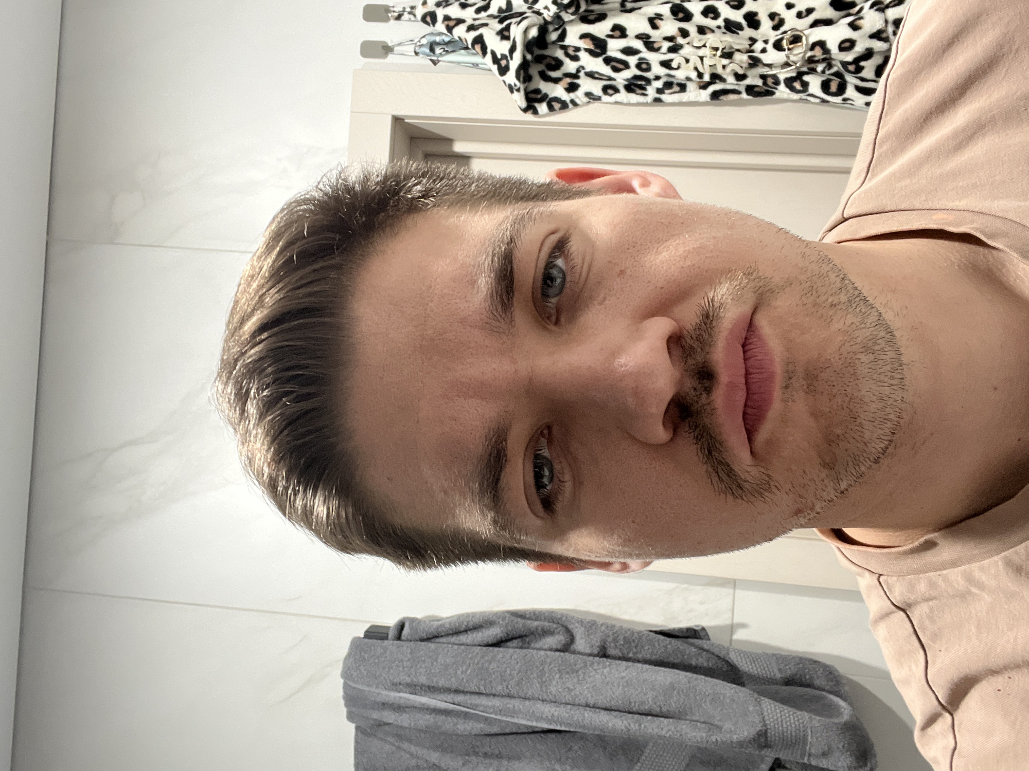 A close-up, selfie-style photograph depicts a light-skinned man with short, brown hair and blue eyes. He has a light beard. He is wearing a light pink shirt. The background shows a white tile wall and a white door with a leopard print garment hanging above it. There is also a gray towel visible. The location in the city of Kazan, Russia, cannot be determined from the available information.