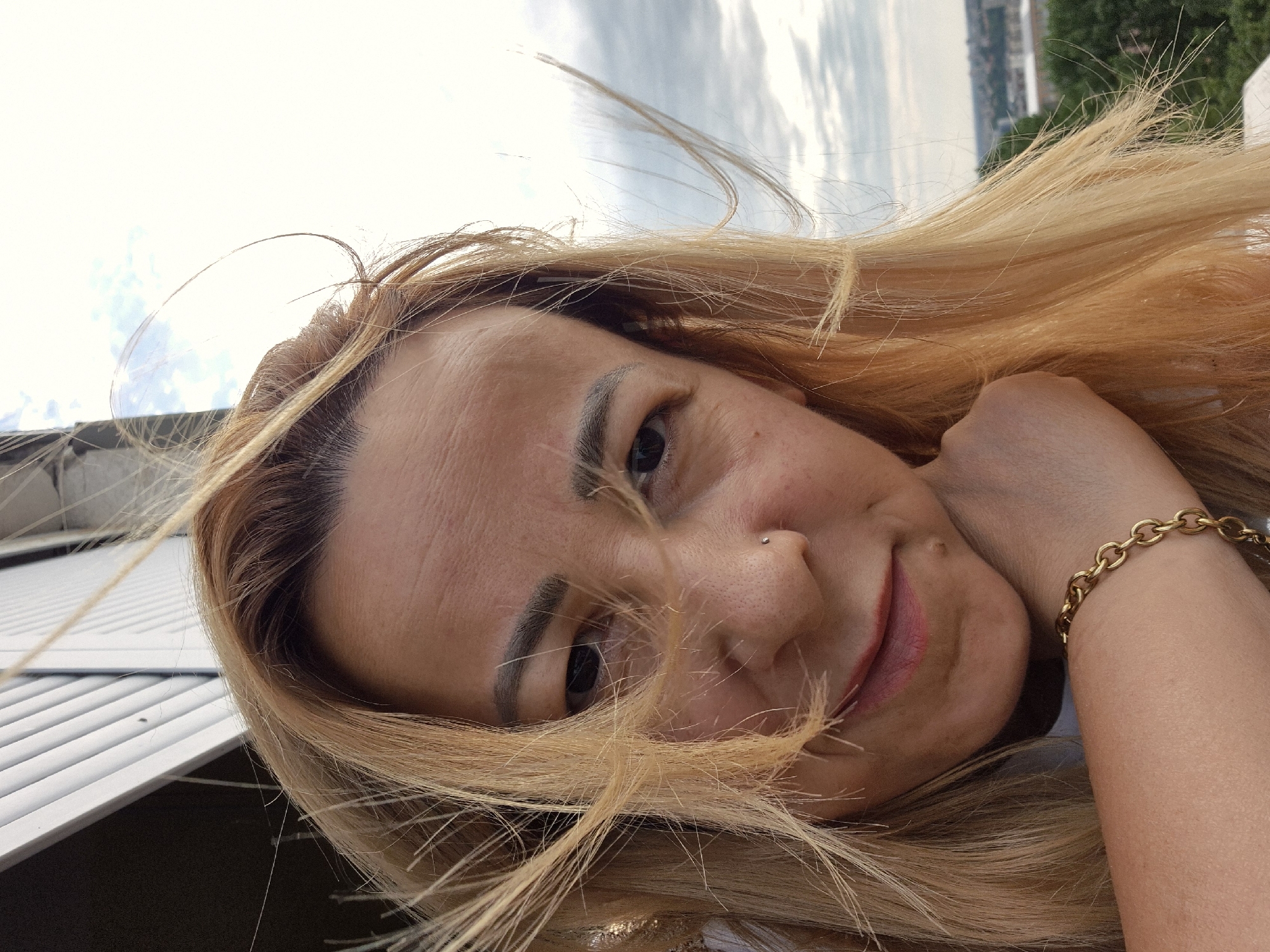 A medium shot captures a blonde woman from the chest up, posing for a selfie outdoors against a backdrop of a cloudy sky and a distant cityscape. Her head is tilted to her left, and her right arm is bent, with her hand resting near her chin. She has a nose ring and wears a gold chain bracelet on her wrist. Her blonde hair is windswept, with strands framing her face and obscuring parts of her forehead and eyes. The woman's skin appears lightly tanned, and she has subtle makeup on her lips and eyebrows. The background shows a hazy view of buildings and trees, suggesting an urban environment.
