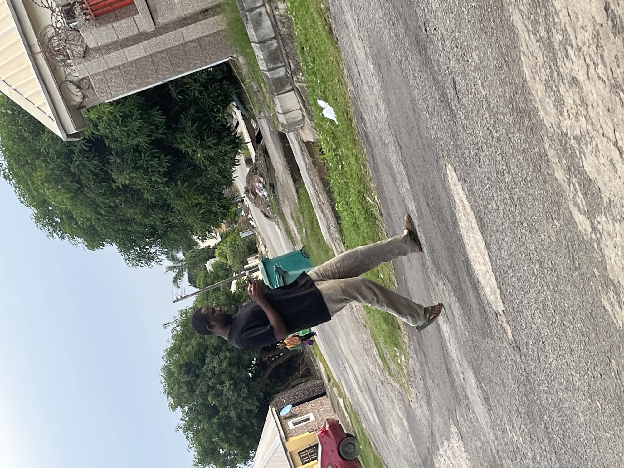 A man in a black t-shirt and beige pants walks on a paved road while looking at his phone. He is wearing sandals. A green trash bin is visible behind him. In the background, there is a building with a sloped roof and barbed wire on top, as well as large trees and a red car. The scene appears to be in a residential area in Abuja, Nigeria, given the architecture and vegetation.