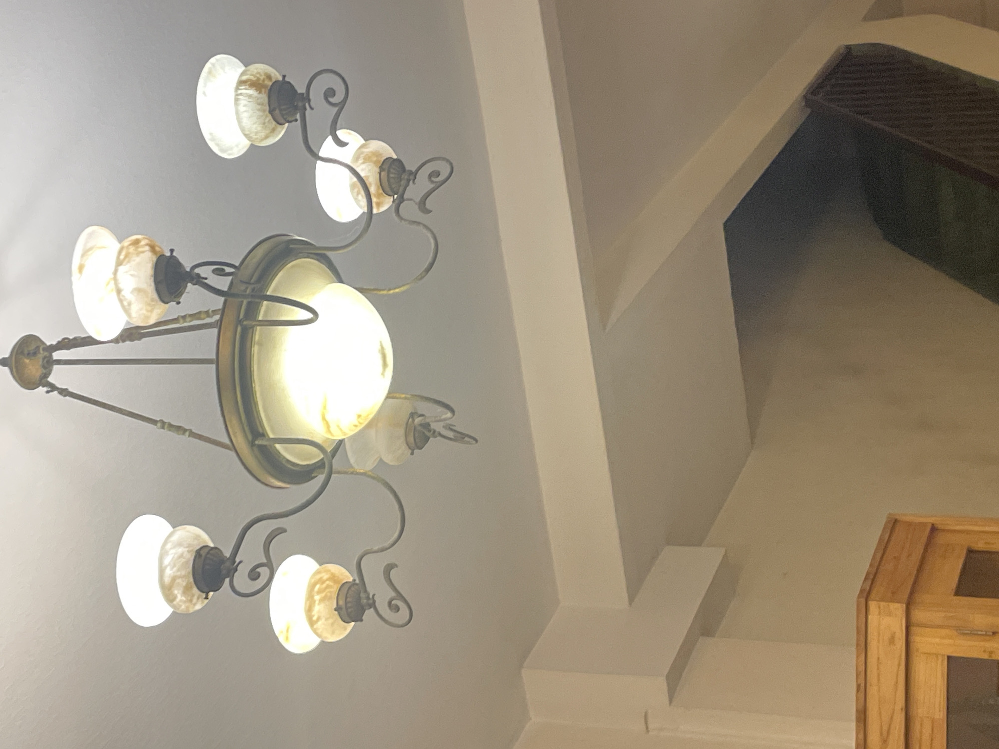 This media file is an indoor photograph taken from a low angle looking upwards towards a ceiling and a chandelier. The primary subject is an ornate chandelier with multiple lights. The chandelier has a metallic frame with decorative scrollwork. It features several glass shades, some of which appear to be made of frosted or marbled glass with warm tones, while others are a brighter white. One central, larger light is prominently illuminated. The chandelier is suspended from the ceiling.
In the background, the ceiling is visible, with angled architectural elements that suggest a sloped or vaulted ceiling. To the right of the chandelier, a portion of a wooden cabinet with glass panes is visible. Further in the background, a dark rectangular grate is set into an opening, possibly a vent or part of a window. The lighting in the scene is warm, emanating from the chandelier itself and creating a soft glow. The overall impression is of an interior space, likely a room within a building. There is no visible text in the image. Given the provided location context of Thủ Đức, Vietnam, the scene likely depicts an interior in that region. The time of day is indeterminate, as the artificial lighting is the dominant light source.