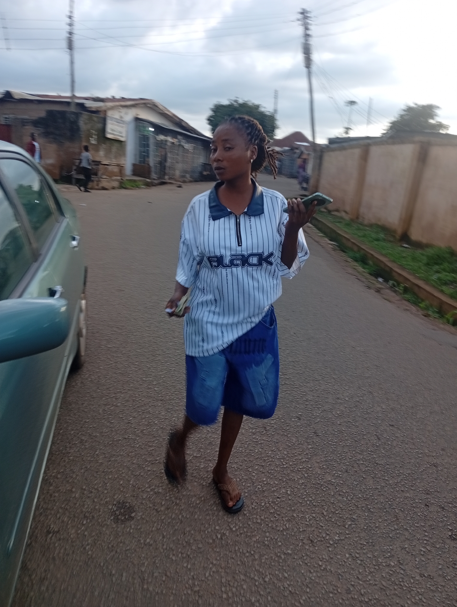A young woman with braided hair walks on an asphalt road in the city of Unknown, Unknown. She is wearing a white and blue striped collared shirt with a zipper and blue denim shorts. In her right hand, she holds a mobile phone, and in her left hand, she clutches a wad of cash. She is wearing brown sandals. To her left, a teal-colored car is parked on the side of the road. The background shows buildings and a wall. The sky is overcast.