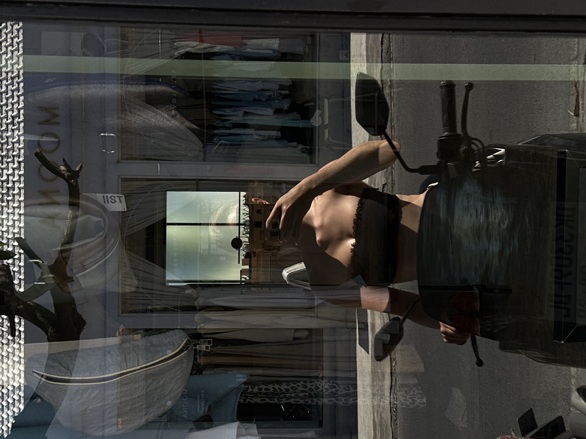 The image, likely taken in North Kuta, Indonesia, shows a complex reflection in a shop window. The reflection captures a woman in a black top seated on a scooter, positioned on what appears to be a sidewalk. The background behind the woman is a neutral gray.
The shop window displays various items, including clothes on hangers, bags, and what appears to be a decorative branch or tree. The shop name, "MOOM", is visible on a sign behind the merchandise. Another sign with the text "IIST" is also visible. These elements combine to create a layered, reflective image, blurring the distinction between the street scene and the store's interior.