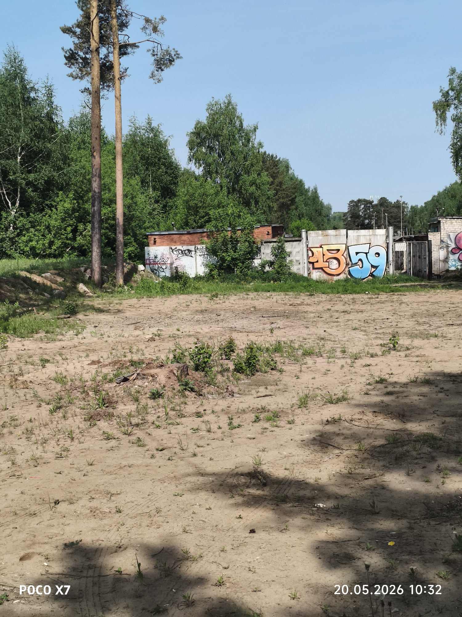 The image shows an empty sandy area near a concrete wall with graffiti in Chaikovsky, Russia. The graffiti includes an orange "45" and a blue "59." Trees are visible in the background. Two tall pine trees are present in the left of the image. There are tire tracks in the sand in the lower part of the image. The picture was taken on May 20, 2026, at 10:32 AM.