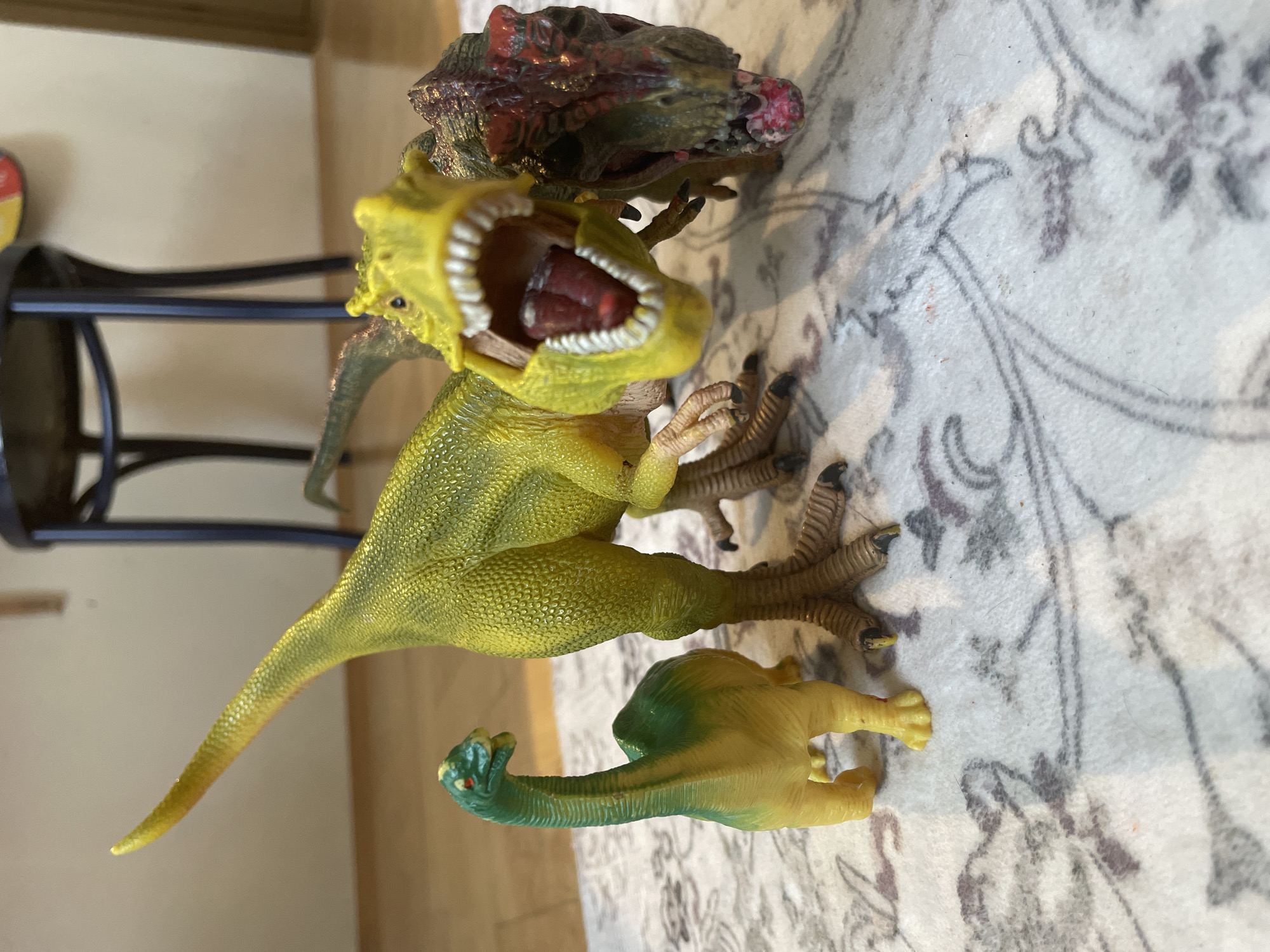 The image shows three dinosaur toys on a patterned rug. A small green and yellow dinosaur with a long neck stands on the left. To its right is a larger yellow dinosaur with its mouth open, showing sharp teeth and a red tongue. Partially behind the yellow dinosaur and to its right is a third dinosaur toy with a reddish-brown head and mouth open. The background is blurred and shows wooden flooring and the legs of a dark chair. The scene is indoors.