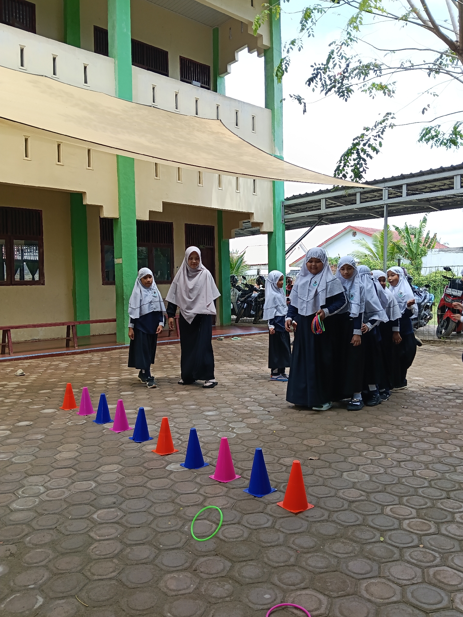 The image shows a group of girls and a woman, presumably students and a teacher, outside a building with green and beige trim. The girls are wearing white headscarves and dark blue uniforms. An adult woman is also wearing a white headscarf, with a larger shawl, and a dark dress.
In the foreground, there is a line of colorful cones and two rings on a paved area with a hexagonal pattern. A small group of girls is standing, some holding rings, as one girl and woman are walking towards the cones.
In the background, there is a building with what appears to be a covered walkway. Several parked motorcycles are visible behind the group of girls. The setting appears to be an outdoor schoolyard, and judging from the environment, this is in Lhokseumawe, Indonesia.