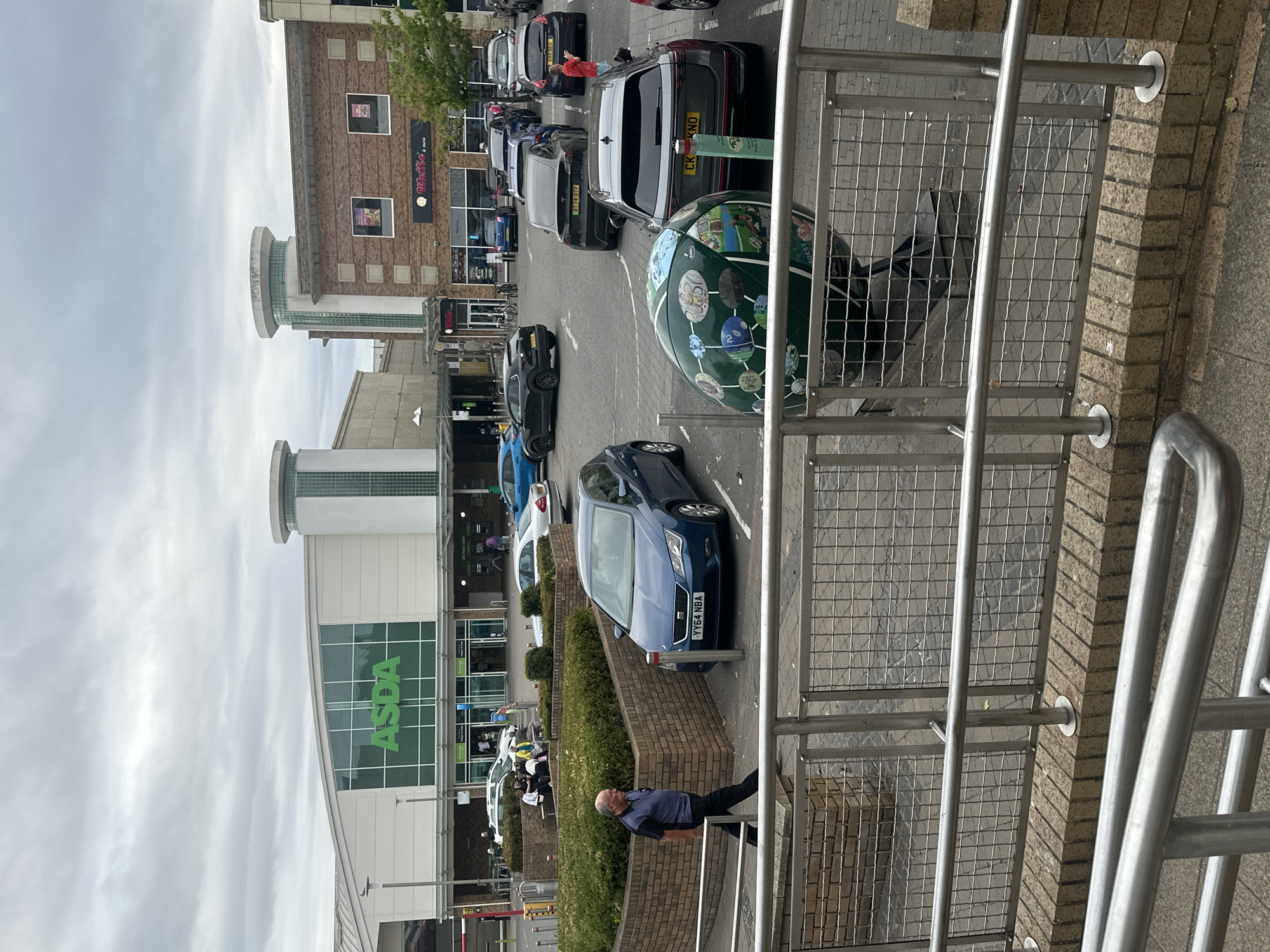 The image shows a view of the Asda supermarket entrance and car park in Rugby, United Kingdom. In the foreground, a man walks past a blue car parked in the car park. Behind him, there is a green, globe-shaped object decorated with rugby-themed imagery. In the midground, shoppers are seen entering and exiting the Asda store, and several other cars are parked in the car park. The background features the Asda building and other shops. The sky is overcast.