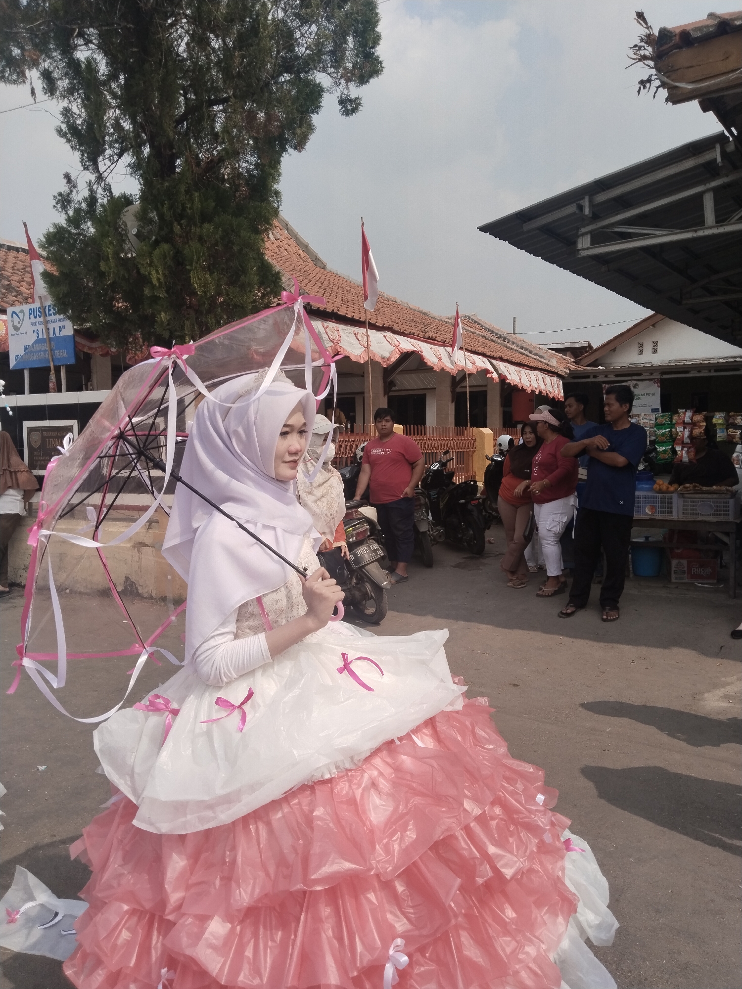 A young woman wearing a white hijab and a puffy white and pink dress, holding a transparent umbrella adorned with pink and white ribbons, stands in a street. The woman is the central focus of the image, and she is looking to her right. Behind her, a group of people are gathered, some standing and talking, while others are near motorcycles. In the background, there are buildings with red tiled roofs and a sign that reads "PUSKESMAS" which is a public health center. Indonesian flags are also visible, indicating a national event or celebration. The scene appears to be outdoors in a semi-urban environment, possibly during the daytime, given the shadows cast on the ground. The presence of the health center sign and local architecture suggests the location is in Margasari, Indonesia.