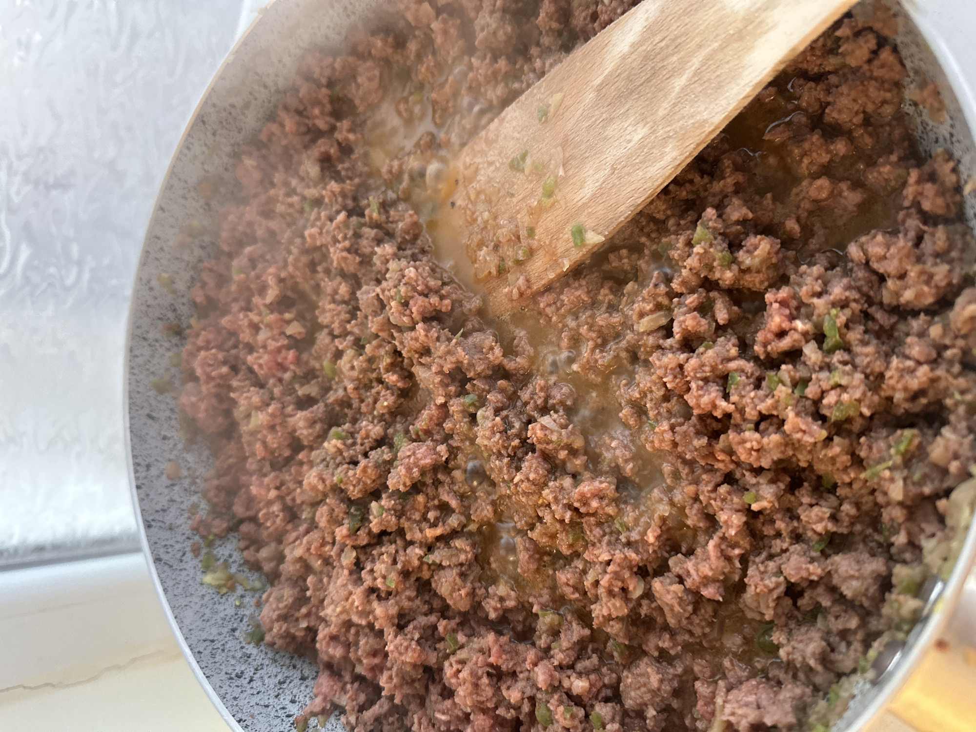 A close-up overhead shot depicts ground meat, possibly beef, cooking in a pan. The meat is browned and mixed with chopped green vegetables, likely peppers. A wooden spatula is present in the pan, stirring or separating the meat. Some liquid, possibly fat or broth, is also visible in the pan. The scene is domestic, suggesting a kitchen environment. There is no information present in the image to determine if this is occurring in Varna, Bulgaria.