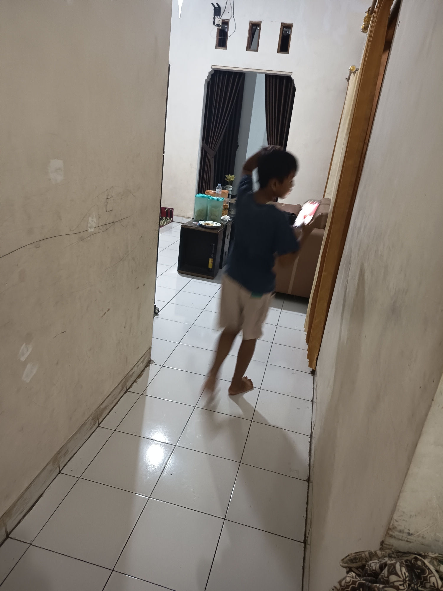 In an indoor environment, a young boy with dark hair, wearing a dark blue t-shirt and light brown shorts, is captured mid-motion as he walks while looking at a lit tablet. His body is slightly blurred, indicating movement. The hallway features tiled flooring and walls, with a wooden door frame visible on the right. In the background, a brown sofa and a black cabinet with a light green container are present.