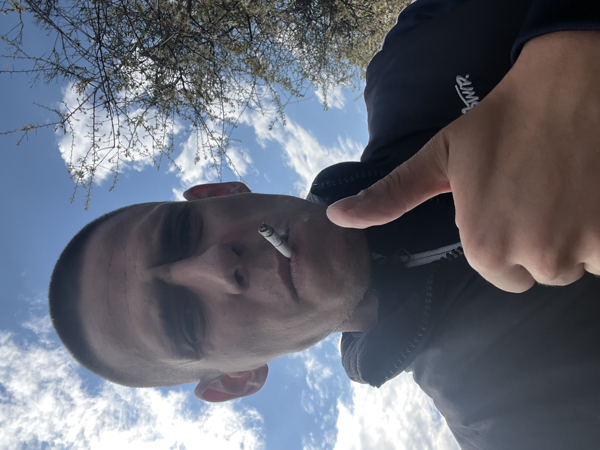 A close-up, low-angle shot of a light-skinned man, with a short buzzcut hairstyle and stubble. He is smoking a cigarette, held in his mouth. He is wearing a dark jacket with a visible white zipper and a logo on the sleeve. His right hand is raised and partially obscuring the view. In the background, a blue sky with scattered white clouds is visible, along with branches from a tree with small white blossoms. The photo may have been taken in Sofia, Bulgaria, but there is nothing in the image to confirm this.