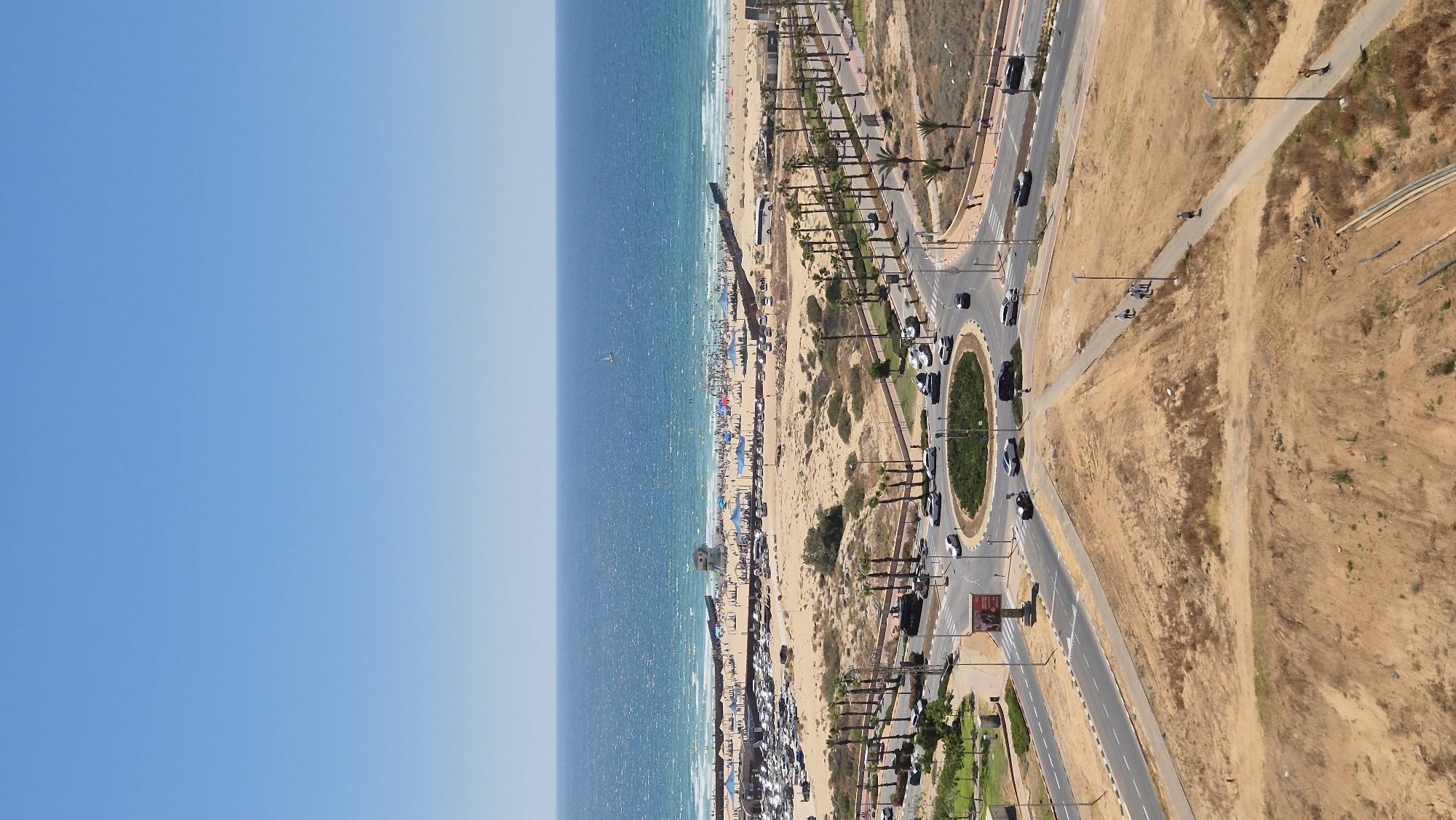 The image captures an aerial view of a beach and adjacent infrastructure in Ashdod, Israel. A sandy beach, crowded with people and umbrellas, lines the coastline next to the blue water of the Mediterranean Sea. A row of palm trees separates the beach from a road where cars are visible. A roundabout with a green center is present on the road. Beyond the road, the terrain transitions to dry, arid land with scattered vegetation. A pedestrian path and additional roads are also visible.