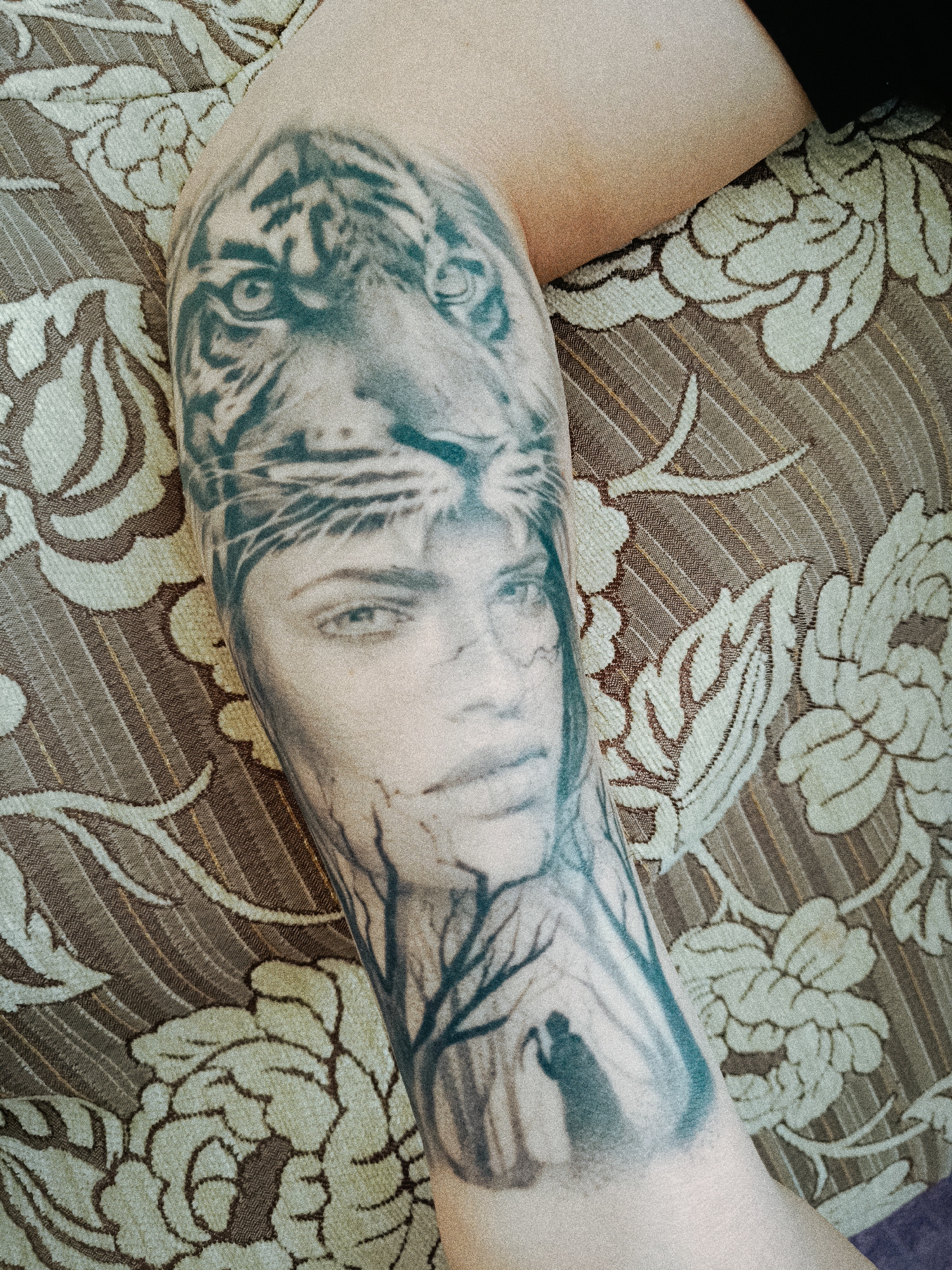 A close-up shot depicts an arm with a tattoo. The tattoo features a combination of images: the head of a tiger, the face of a woman, a forest scene with bare trees, and a figure resembling the Grim Reaper. The arm is resting on a patterned fabric surface. No information connects the image to Tobolsk, Russia.