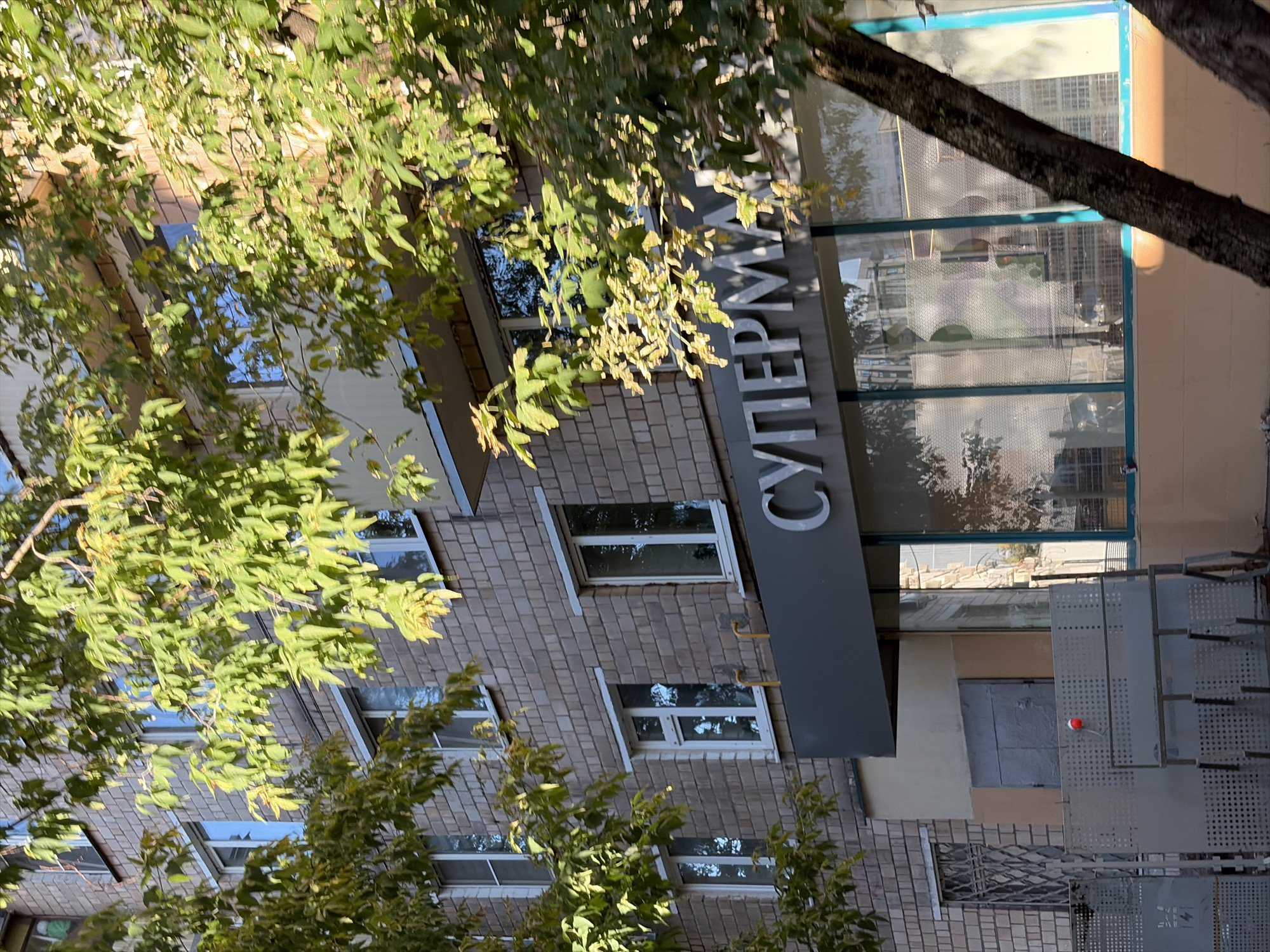 A brick apartment building in Moscow, Russia, is partially obscured by the branches of a tree with green and yellowing leaves. The building has multiple windows with white frames and yellow pipes visible on the facade. A dark gray sign with large silver letters spelling "СУПЕРМАРКЕТ" (supermarket) is mounted above a storefront with large windows. The storefront windows reflect the surrounding environment, including other buildings and trees. In front of the building, there are metal fences and a gray metal door. A small red object is attached to one of the fences. The image is taken from a low angle, looking up at the building.
