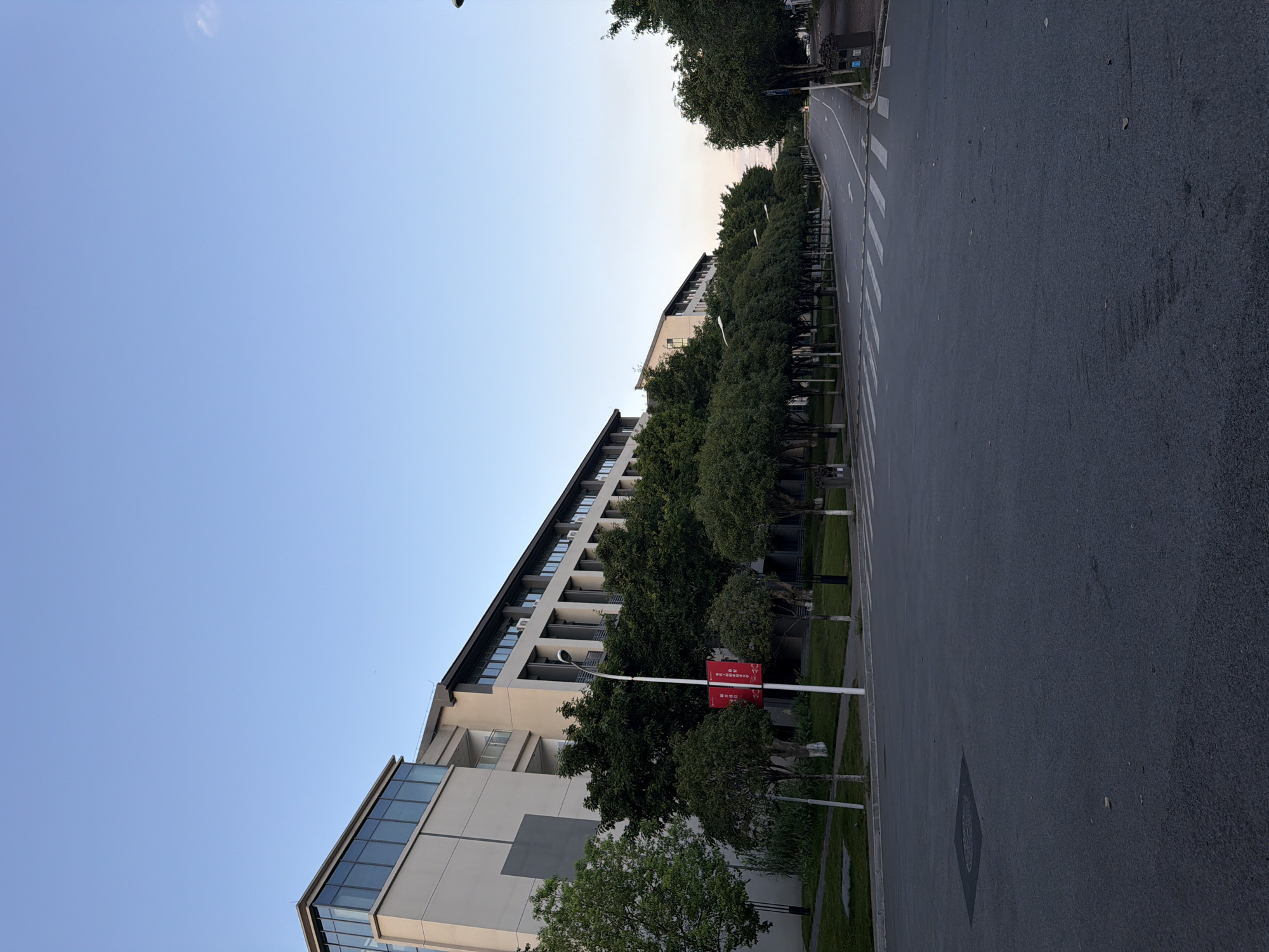 This is an outdoor, eye-level, full shot of a street in Jiangjin, China. The street is lined with trees and a modern, multi-story building with a glass-fronted section on the left. The sky is clear and blue. The road is asphalt with white crosswalk markings. There is a lamppost with a red banner on the left side of the road. There are no people visible in the image. The environment appears to be quiet and orderly.