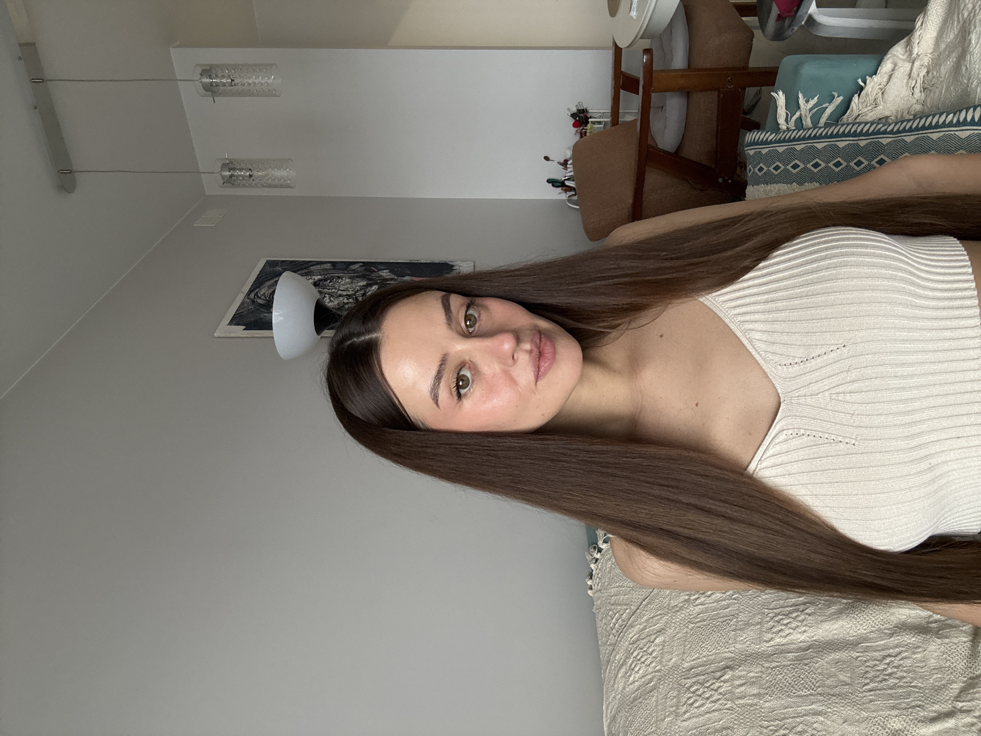 The image shows a woman with long, brown hair lying on a textured, light-colored bedspread. She is wearing a white, ribbed tank top. Behind her are neutral-colored walls, a picture on the wall, and a modern-style ceiling light fixture. In the background, there are chairs and a small table with some objects on it. There's not enough information in this image to determine if this scene is located in Krasnoyarsk, Russia.