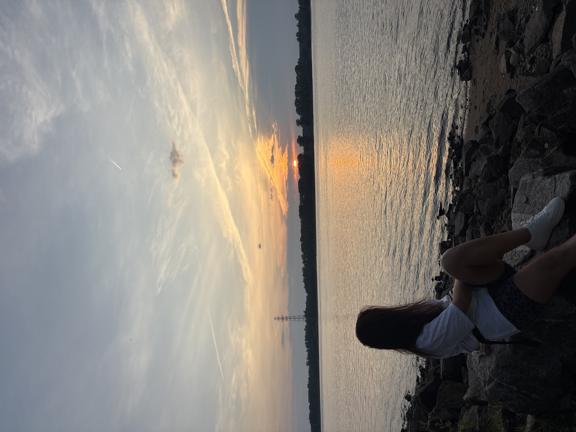 A young woman is sitting on rocks by the water, facing away from the camera and looking at a sunset over a body of water. The sky is a mix of blue and orange hues, with clouds scattered throughout. A tall transmission tower is visible in the distance, along with a silhouetted treeline on the opposite shore. The water is calm, with gentle ripples reflecting the colors of the sunset. The woman is wearing a white t-shirt, patterned shorts, and white sneakers. The scene appears to be a peaceful and scenic overlook. This location is the embankment of the Kama River in Chaikovsky, Russia.