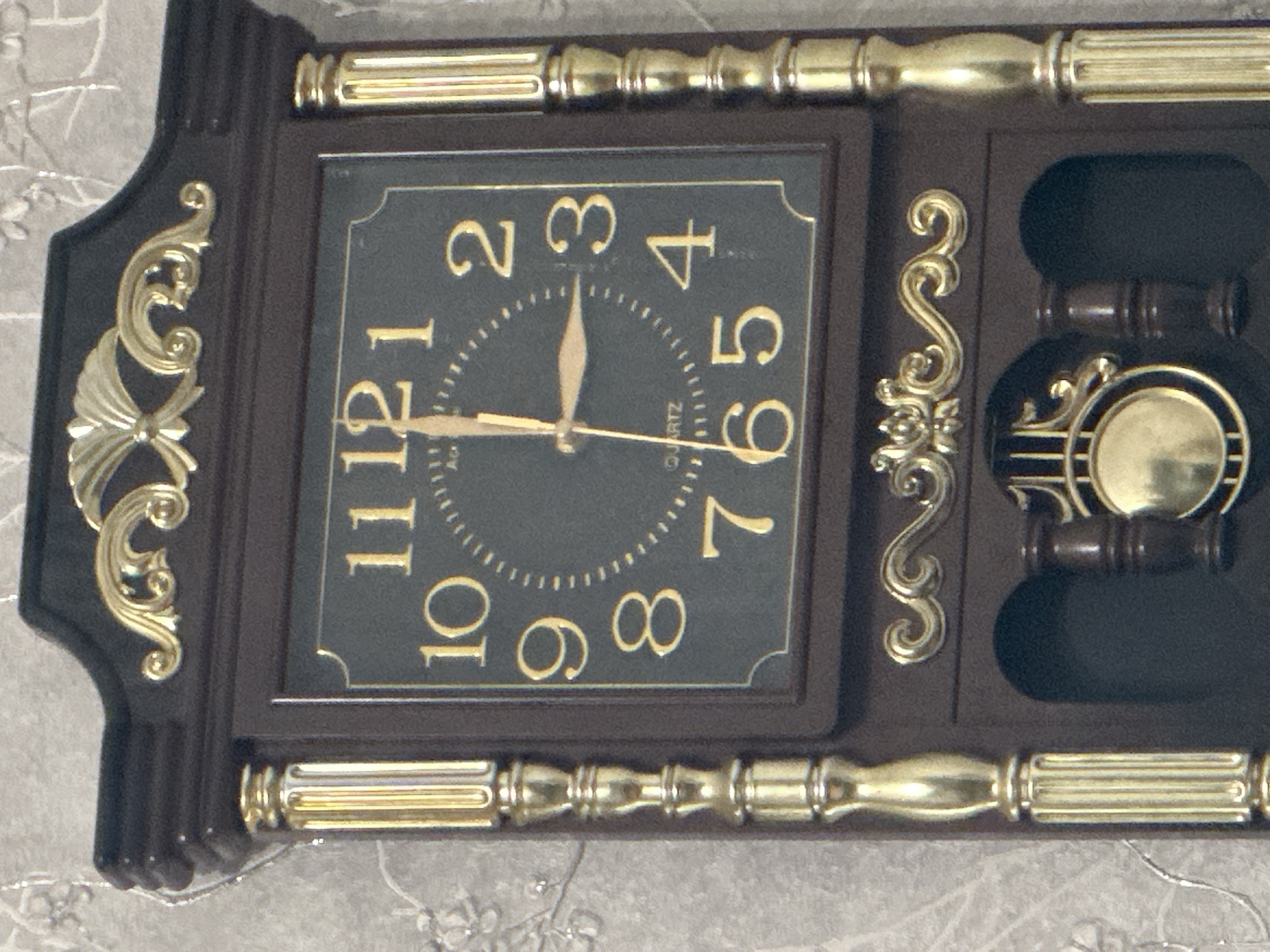 The image displays a close-up, tilted view of a decorative wall clock, likely situated indoors in Minsk, Belarus. The clock features a dark brown or black rectangular frame, richly embellished with gold-colored decorative elements. Ornate gold scrollwork adorns the left and right sides of the central clock face, and matching gold horizontal bars with fluted and spherical patterns are positioned above and below the clock face, as well as above and below a decorative panel on the right side.
The clock face itself is a dark gray or black, featuring prominent gold Arabic numerals from 1 to 12. A circular minute track with small gold dashes is visible around the center. The hour hand points between 11 and 12, slightly closer to 11, while the minute hand points directly at the 6, indicating the time is approximately 11:30. Small, vertically oriented text reading "QUARTZ" is visible near the bottom center of the clock face. A small, blurry inscription, possibly "Ac", is also present near the center. To the right of the main clock face, a separate compartment within the casing contains a decorative element that resembles a pendulum or chime, also featuring gold and dark accents. The clock is set against a light-colored wall with a subtle, light gray patterned wallpaper or fabric, hinting at a delicate floral or abstract design. The clock's design evokes a traditional or classic aesthetic.