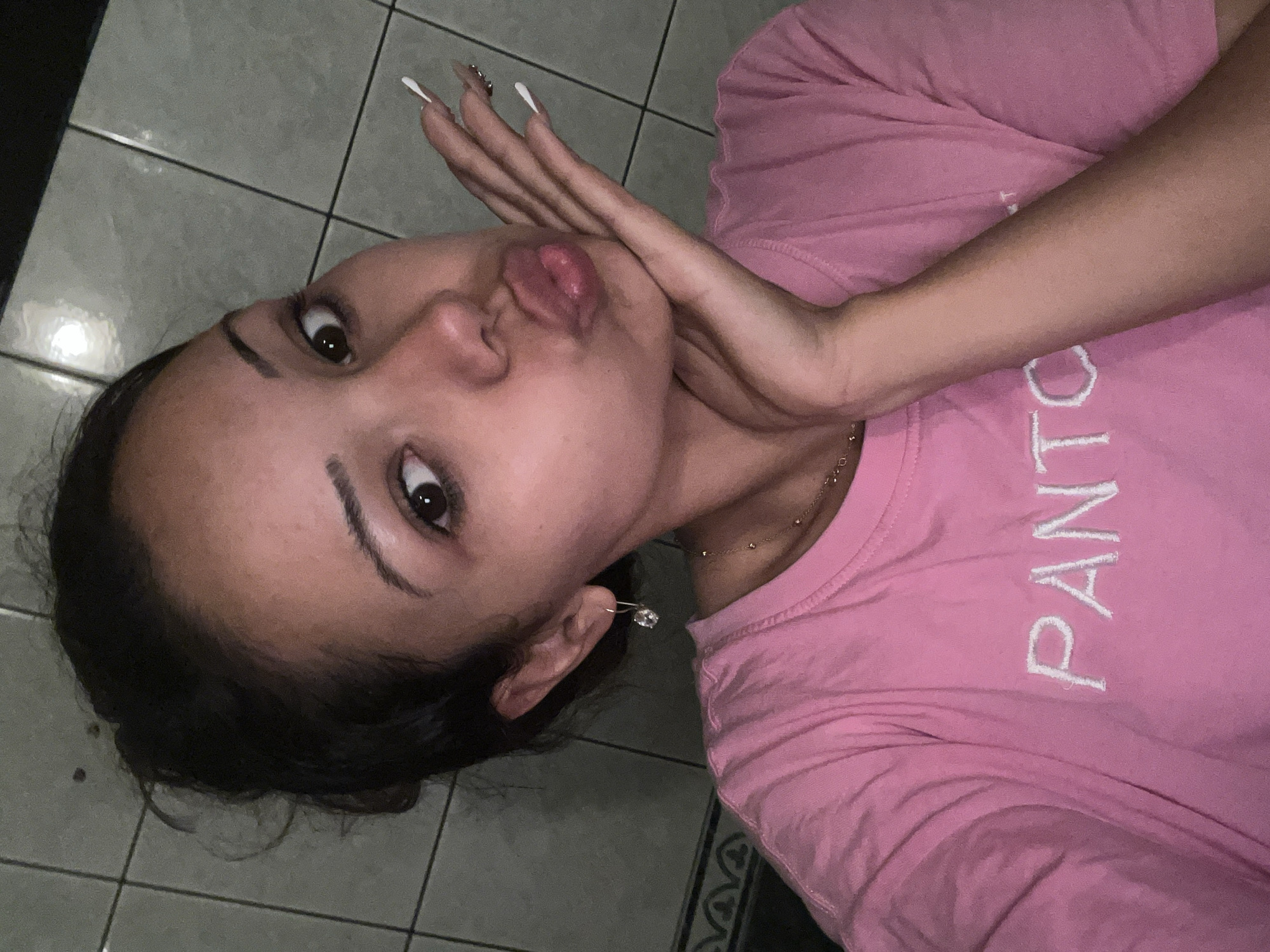 A young adult female is depicted in a medium-shot selfie, posing with pursed lips and wide, dark eyes looking directly at the camera. Her dark hair is pulled back, with some loose strands framing her forehead. She is wearing a short-sleeved pink t-shirt with the white embroidered text "PANTO" visible on the chest. A thin gold-colored chain necklace is visible around her neck. A dangling, clear faceted earring is present in her left ear (viewer's right). Her right hand (viewer's left) is raised, fingers splayed and curved, with the palm resting against her chin and jawline. Her fingernails are long, manicured, and feature white tips. The background consists of light gray square tiles with dark grout lines, suggesting an indoor tiled wall. A decorative tiled border is visible at the lower left. The lighting is artificial and appears somewhat dim. The location is Unknown, Unknown.