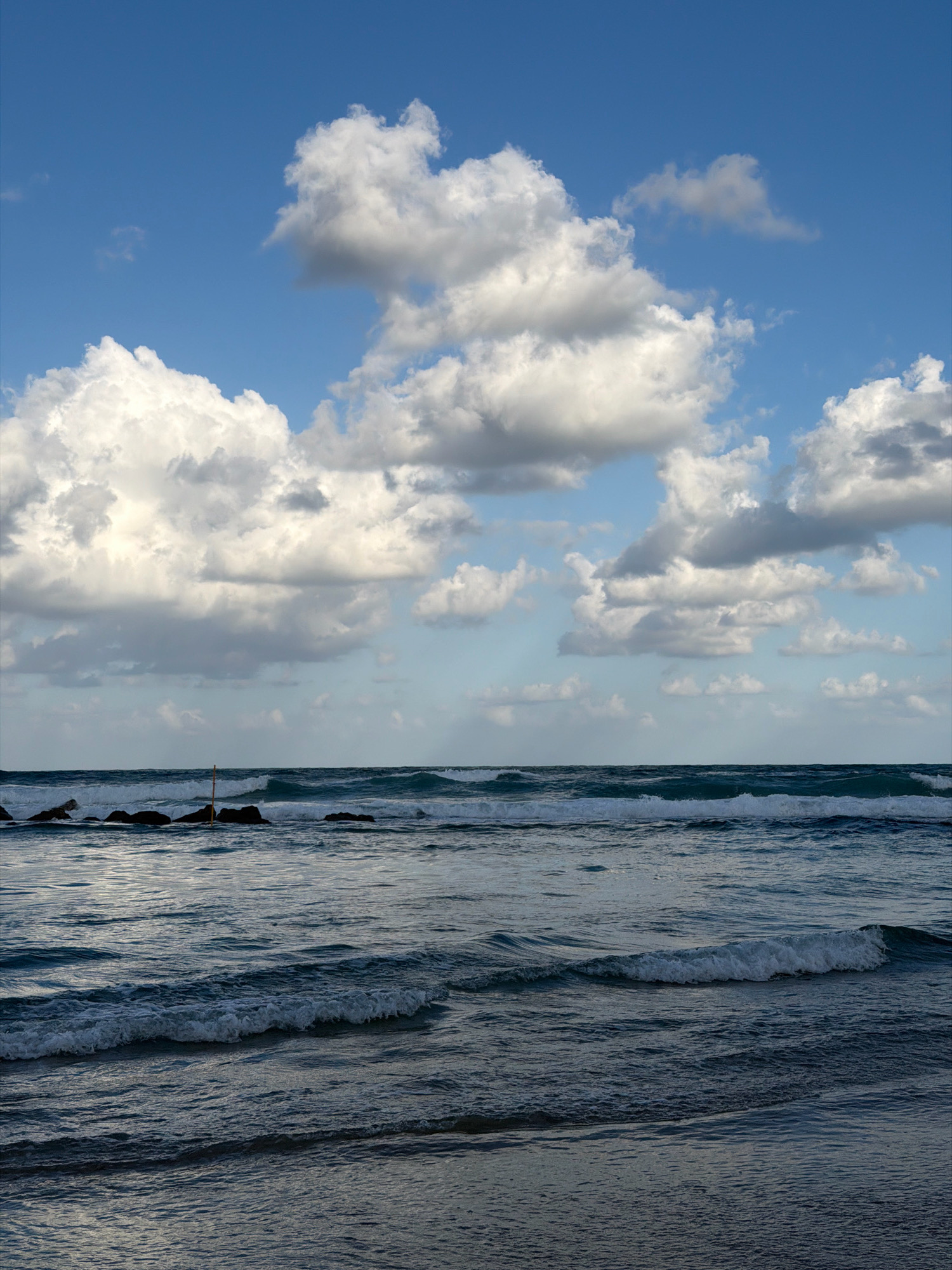 The image captures a view of the sea from the coast of Bat Yam, Israel. The sky is a vibrant blue, dotted with fluffy white clouds. The sea below is a deep, dark blue with white-capped waves rolling towards the shore. In the foreground, the waves are more defined, showing the texture and movement of the water. Towards the middle ground, a line of dark, rocky outcrops emerges from the sea, with a single yellow pole planted in the water amongst them. The scene is serene and natural, with no people or man-made structures other than the pole. The lighting suggests it is daytime, possibly late afternoon, as indicated by the soft light and shadows on the water.