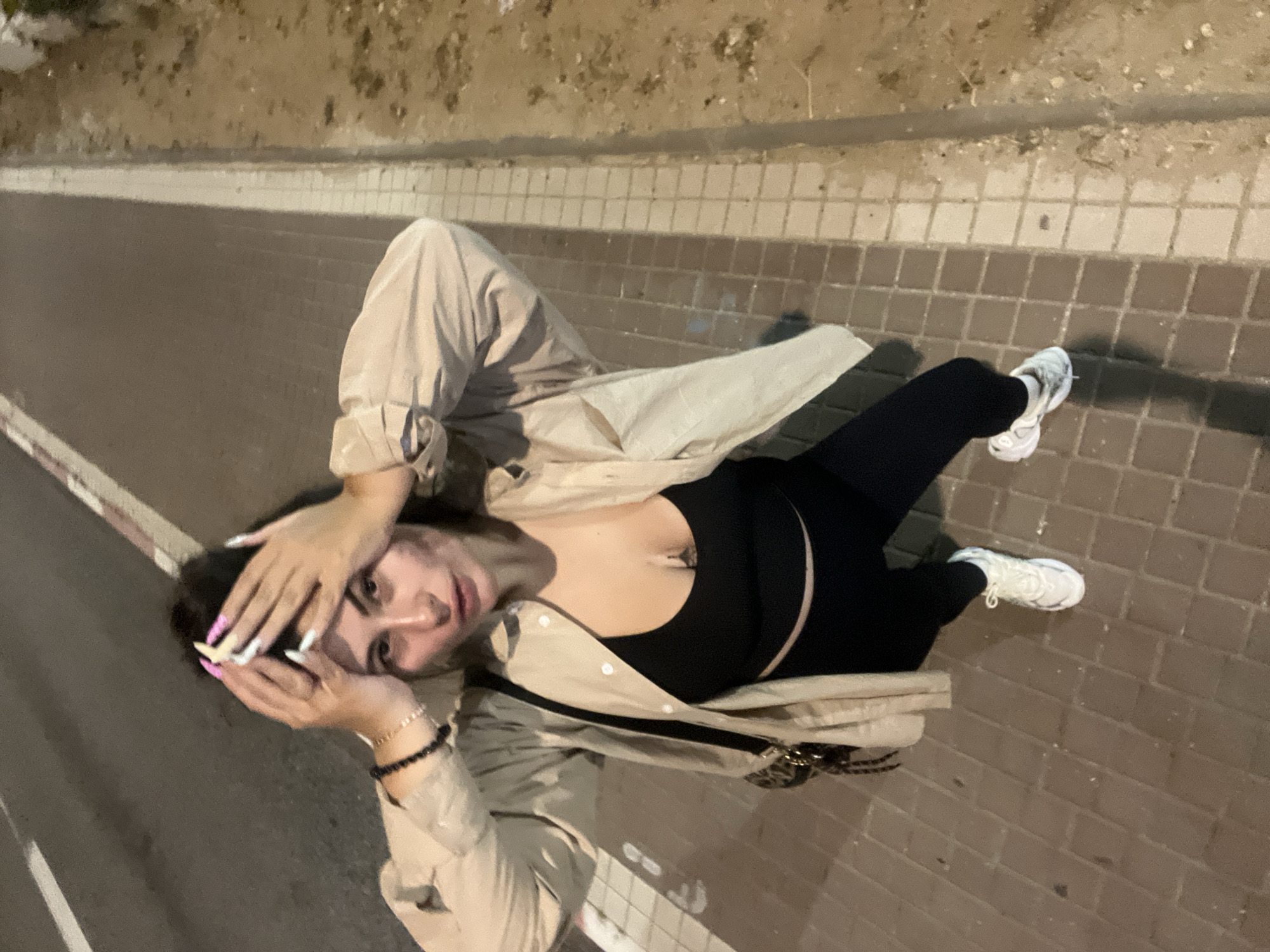 A high-angle shot shows a woman lying on a brick walkway next to a curb and sidewalk. The scene appears to be at night. The woman has on a black top, black leggings, and white sneakers. She is wearing a long beige jacket over her outfit. Her hands are raised near her face. A black beaded bracelet is visible on her left wrist.