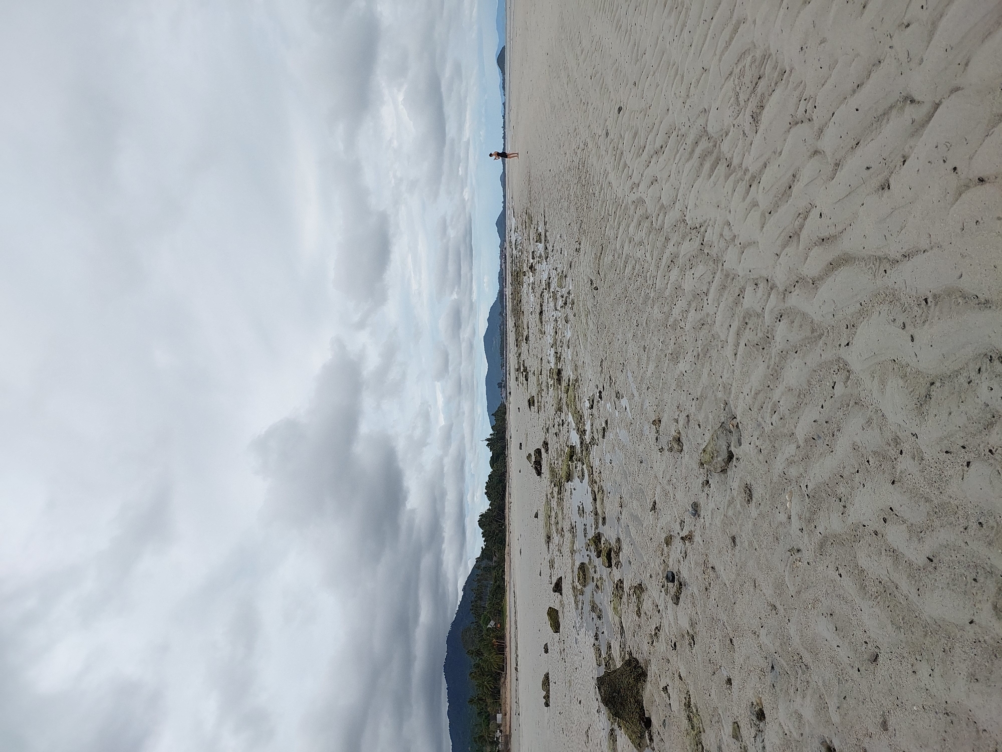 A wide, sandy beach stretches into the distance under a heavily overcast sky. The sand is marked with the rhythmic patterns of receding tides, and scattered rocks and shells litter the foreground. In the middle ground, a lone figure stands near the water's edge, facing away from the camera. Beyond the beach, a line of lush green trees and palm trees can be seen, backed by a distant, hazy mountain range. The overall impression is one of a quiet, natural landscape, possibly during low tide. The location is identified as Ko Samui District, Thailand.