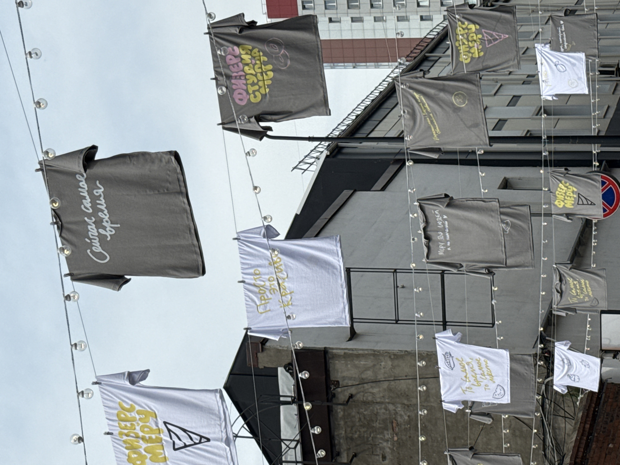 In an outdoor urban setting, multiple t-shirts are displayed on a clothesline strung between buildings under a cloudy sky. The t-shirts are printed with various text and graphics, some in Cyrillic script. Some of the t-shirts feature ice cream cone designs and phrases like "Физика", "Меру", "Это самое время", and "Просто это красиво". String lights are also hung on the same line as the t-shirts. The background includes architectural elements of buildings, including brickwork, windows, and rooftop structures.