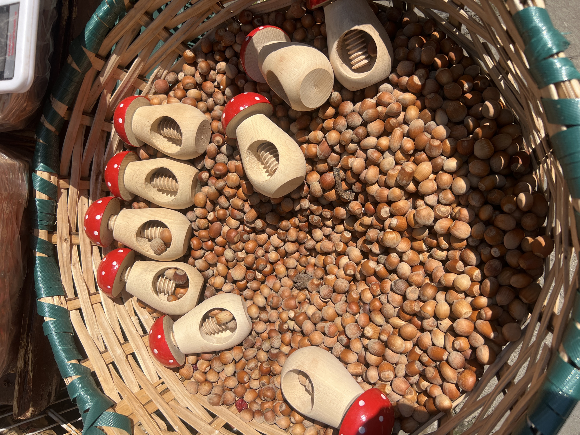 In Yaremche, Ukraine, a large woven basket overflows with raw hazelnuts. Scattered among the nuts are several wooden nutcrackers shaped like red-capped mushrooms. The nutcrackers are light-colored wood with red tops decorated with white dots, resembling toadstools. Each nutcracker has a visible metal screw mechanism designed to crack nuts. The basket is lined with a green woven material. The scene appears to be an outdoor market setting, possibly for sale, given the arrangement and presentation. Sunlight casts shadows across the nuts and basket, indicating it is daytime.