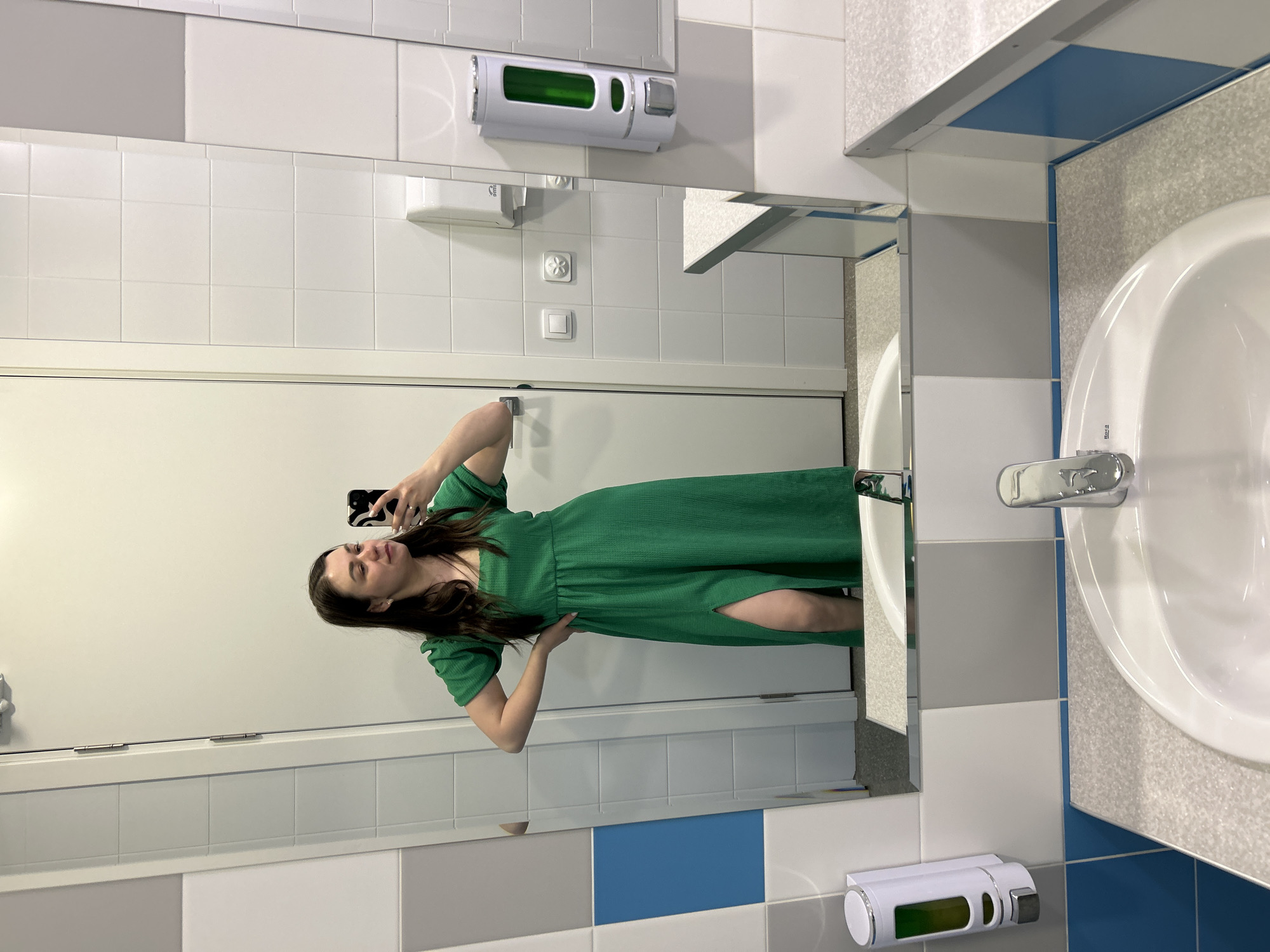 A woman is taking a selfie in a bathroom. The bathroom features a sink, mirror, soap dispenser, hand towel dispenser, and tiled walls in white, blue, and grey. She is wearing a green dress with a slit. The location is possibly Izhevsk, Russia.