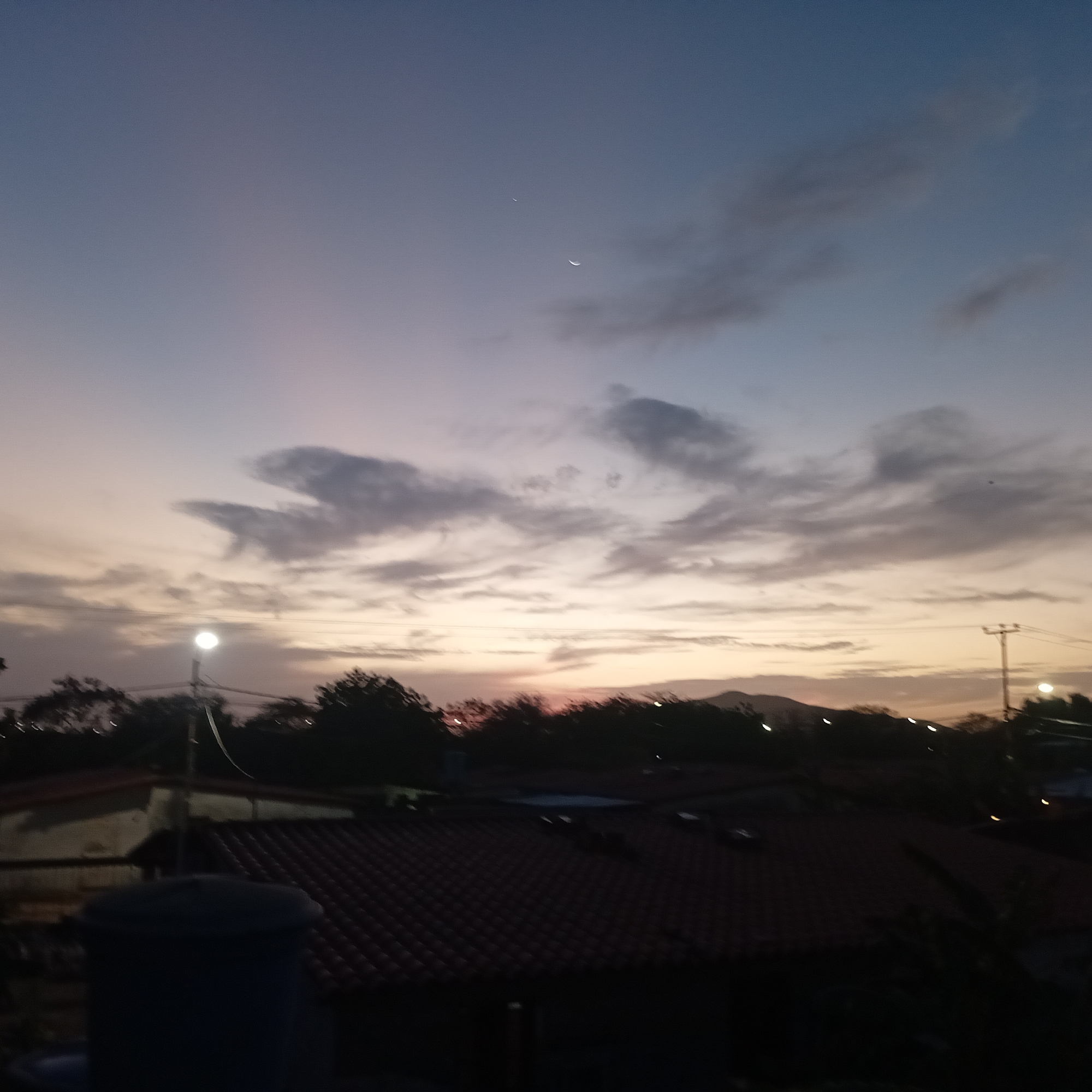 The image shows a twilight scene in an urban area. The sky is a gradient of blue and pink hues, with scattered clouds. A crescent moon and a star are visible in the sky. Below, rooftops of houses with red tile roofs are visible. In the midground, there is a line of trees and a utility pole with wires. A water tank is visible in the foreground. The location is Unknown, Unknown. There are no people visible.