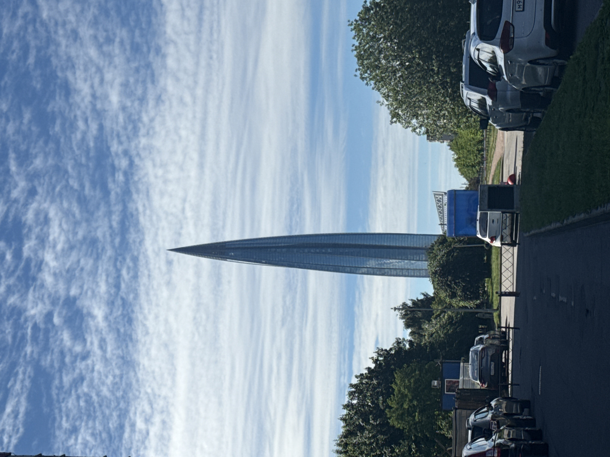 The image depicts the Lakhta Center skyscraper, a prominent landmark in Saint Petersburg, Russia, on a sunny day with scattered clouds. The skyscraper, characterized by its distinctive needle-like shape and glass facade, is the central focus of the image. In the foreground, a paved road leads towards the skyscraper, with several cars parked along the right side of the road and more cars on the left. A barrier system, likely for controlling vehicle access, is visible across the road. Lush green trees and vegetation flank the sides of the road, and the overall environment suggests an urban setting on the outskirts of the city, possibly near a business or industrial complex given the presence of portable structures and fencing. The sky is a clear blue, with wispy white clouds forming patterns across it. There are no visible people actively engaged in any specific actions within the frame.