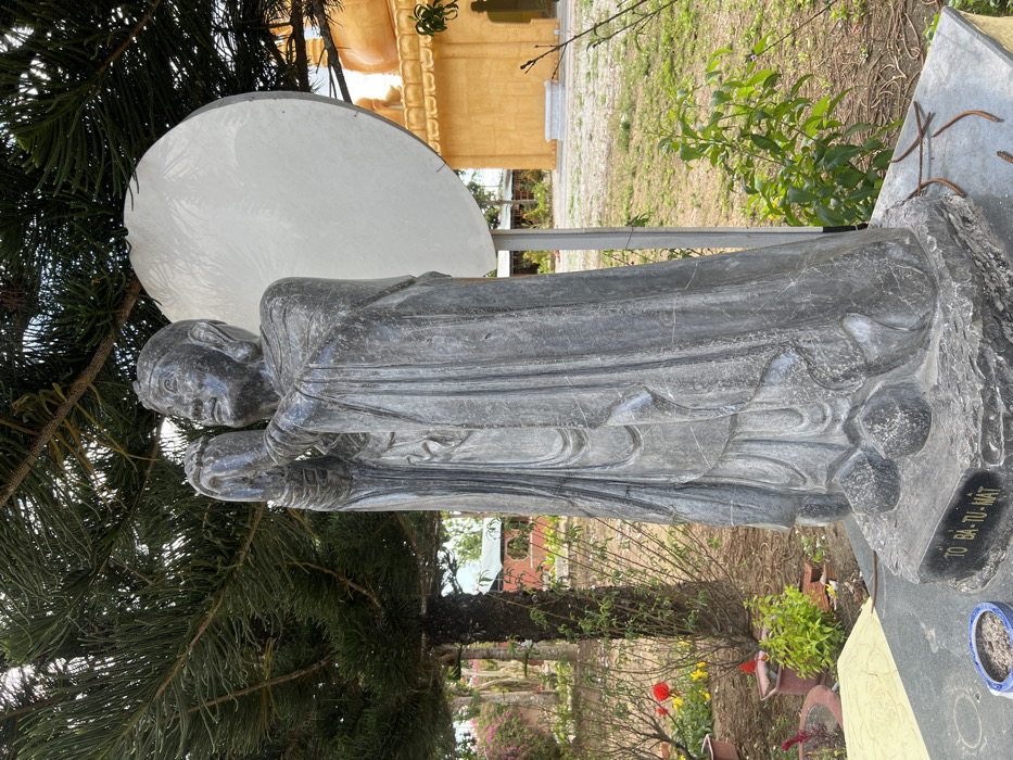 An outdoor scene in Đăk Mar, Vietnam, depicts a dark grey stone statue of a robed figure positioned centrally. The figure stands with hands clasped together at chest level, looking slightly upwards, and wears a cap-like head covering. The statue rests on a dark grey stone base, which bears the inscription "TO BA-TU-MAT" on its front edge. A small, partially visible blue bowl containing dark material sits on the base to the left of the inscription.
Behind the statue, a large pine tree with green needles extends into the upper left quadrant of the frame. To the left of the statue, a garden area features red and yellow flowers, various green plants, and potted plants, with smaller red-roofed buildings visible in the distance. To the right of the statue, a large, round, white panel or disk stands upright, partially obscuring a prominent golden temple building with multi-tiered, curved roof details. The ground around the statue is a mix of sparse green grass and bare earth, with fallen pine needles or small branches scattered on the statue's base and the ground. The scene is illuminated by natural daylight. No individuals are present in the image.