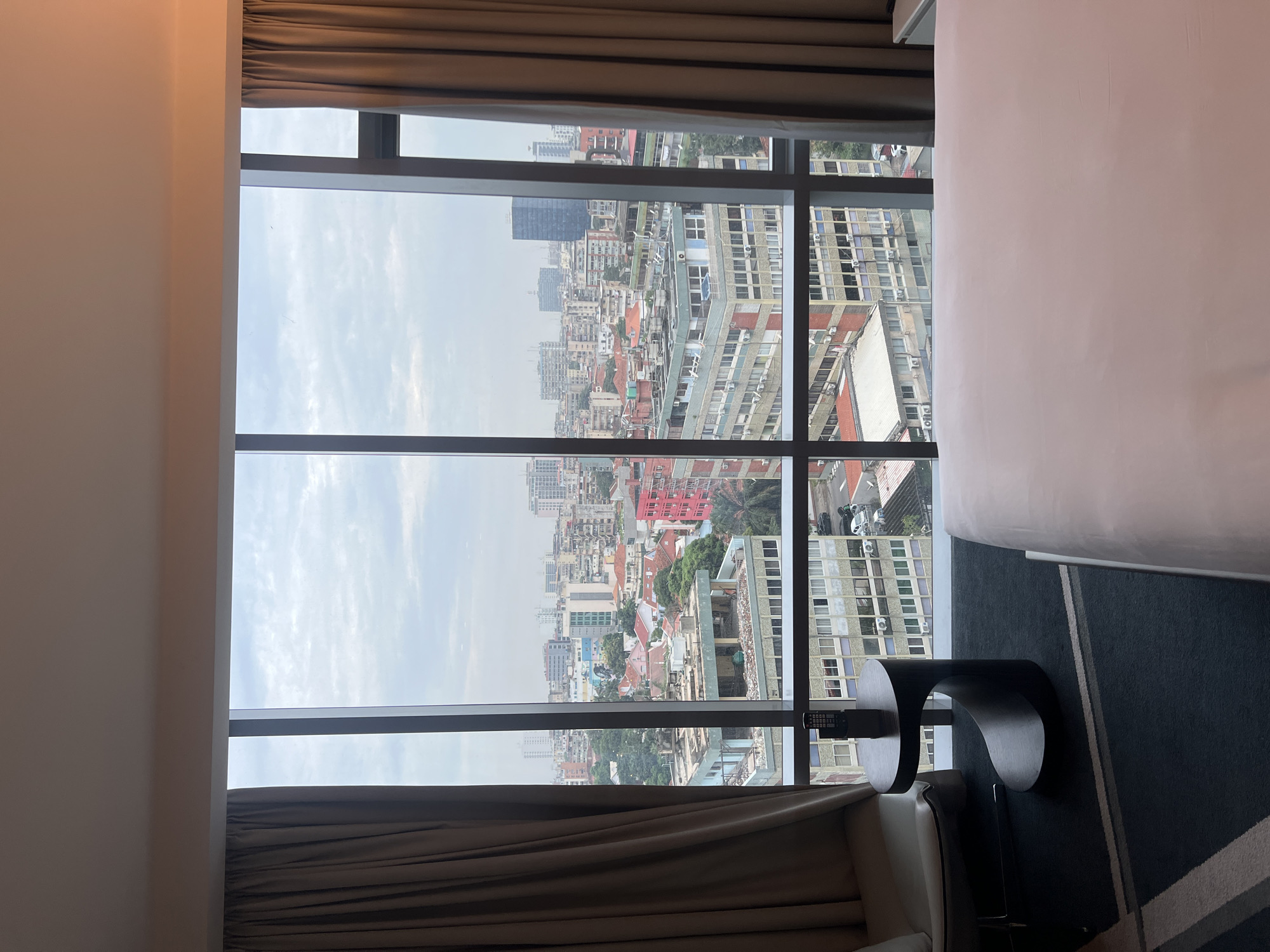 A vertical view from inside a room with a floor-to-ceiling multi-pane window, overlooking a dense urban landscape. The sky is overcast, casting a soft light on the cityscape. Buildings vary in height, architecture, and color, with a mix of high-rise structures and lower-rise buildings clustered together. In the foreground, the room has neutral-colored curtains partially framing the window. A section of a bed is visible on the right side of the frame, and modern furniture, including a small side table and chair, is also present in the foreground. The scene suggests a hotel room or apartment with a view of a bustling city.