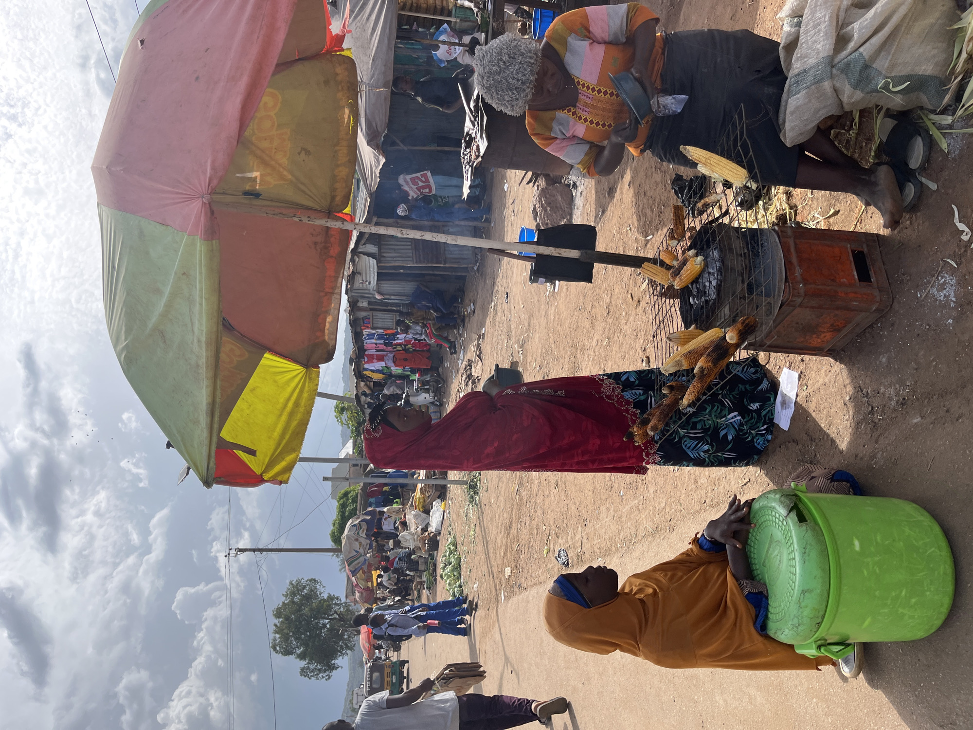 This image depicts an outdoor market scene in Jos, Nigeria. The focus is on several individuals interacting around food stalls. In the foreground, a person wearing an orange headscarf and matching dress is seated next to a large green plastic container, resting their hands on its lid. To their left, another person in a red headscarf and dress is standing, facing away from the camera, with some corn cobs visible near them.
Behind these individuals, a woman with grey hair, wearing an orange and patterned top, is seen tending to a makeshift grill where corn is being roasted. She appears to be engaged in preparing or selling the corn. The grill itself is placed on top of a rusty orange metal structure, with several cobs of corn cooking over coals. An umbrella, with alternating pink, yellow, and green panels, provides shade over a portion of the market area.
The background reveals more of the market with various stalls and people. In the distance, there are displays of colorful clothing and produce. The ground is dusty and unpaved, typical of many market settings in the region. The sky above is partly cloudy, suggesting it is daytime, likely late morning or afternoon, given the lighting. The overall atmosphere appears to be that of a busy, functioning marketplace. There is no visible text in the image.