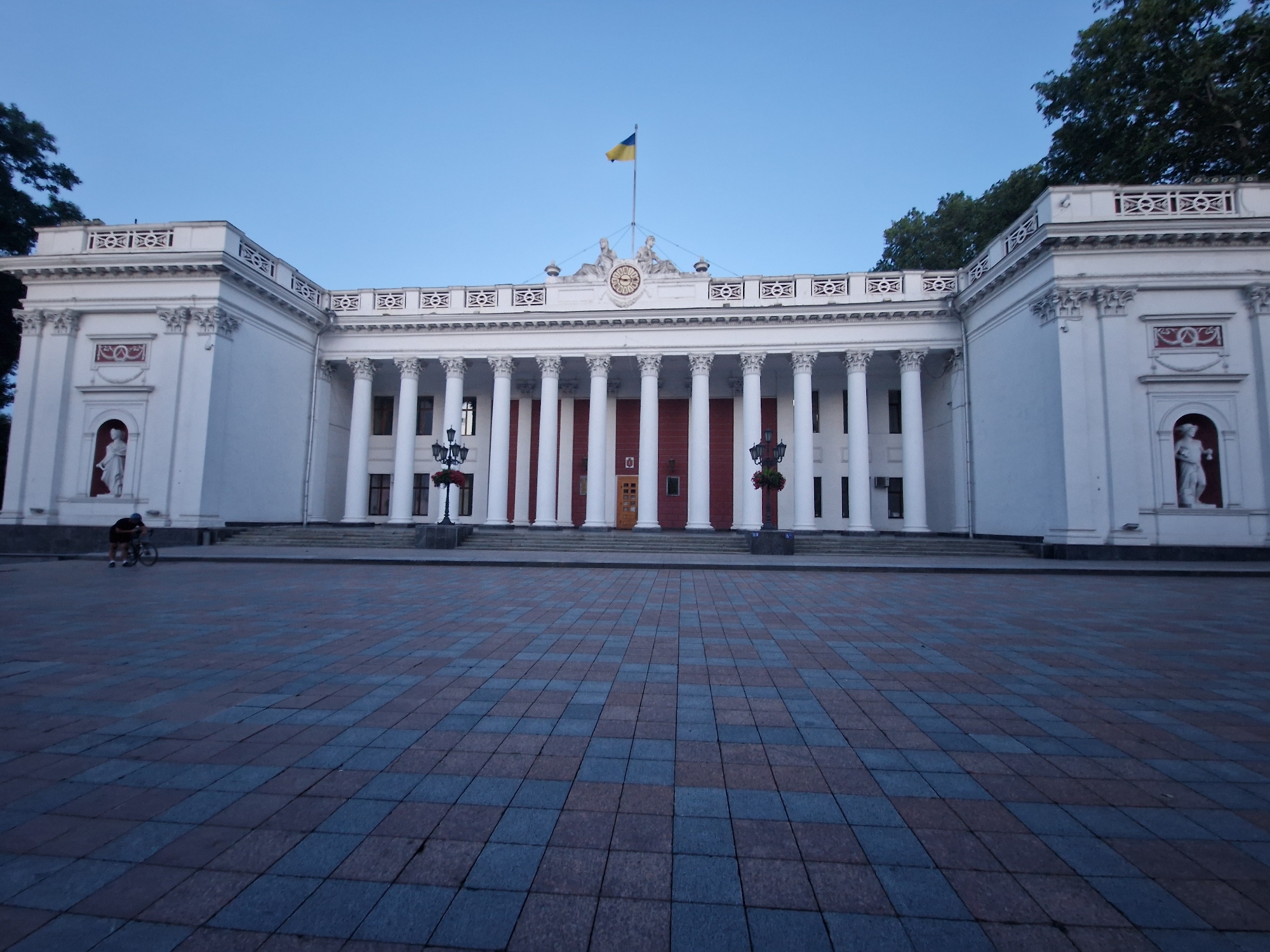 This is a wide-angle photograph taken from a low perspective of the facade of a large, white, neoclassical building. The building features a prominent colonnade of tall, white pillars, with a red facade behind them. The facade is topped by a white entablature with decorative friezes and statues. A Ukrainian flag flies from a flagpole centered above the entablature. A clock face is visible on the building's facade, directly below the flag.
In front of the building is a large, paved plaza with a checkered pattern of blue and reddish-brown tiles. To the left of the plaza, a person is riding a bicycle. The scene is set under a clear, light blue sky, suggesting it is early morning or late afternoon due to the soft lighting and lack of harsh shadows. The overall mood of the image is serene and architectural. There is no visible text in the image.