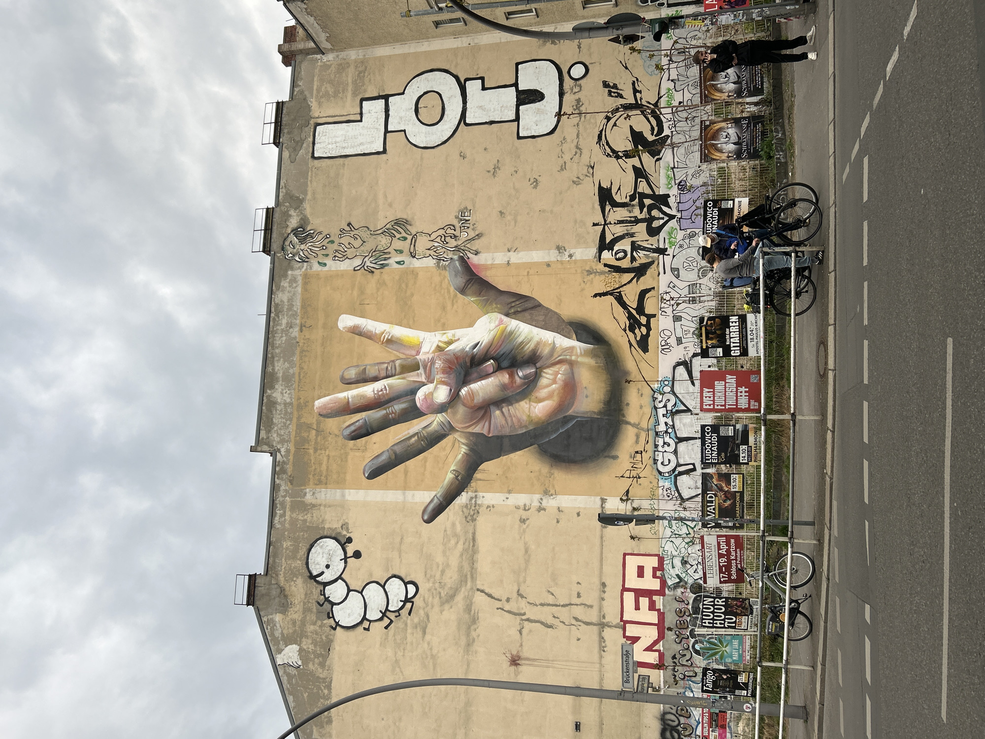 This media file captures an urban street scene in Berlin, Germany, featuring a large building wall adorned with an expansive mural and layers of graffiti. The focal point of the mural is a pair of realistic hands, seemingly emerging from the wall, with one hand making a peace sign (two fingers raised) and the other showing a three-finger gesture. Above the hands, a large, stylized white graffiti tag reads "CHOP", and various black abstract tags are scattered across the wall. Below the hands, a whimsical white cartoon caterpillar character is depicted, and to the left, two smaller, tentacled creatures are drawn.
The building wall itself is a weathered, light beige color, heavily covered with an array of concert and event posters, advertisements, and numerous graffiti tags. A metal fence bordering the sidewalk is also plastered with these flyers. Along the street, which is asphalt with white dashed lane markings, several people are present. One person stands near the fence, seemingly using a phone, while two others are on bicycles, one appearing to be riding. The sky is overcast, indicating a daytime setting without direct sunlight.
Visible text on the wall and posters includes "CHOP" (graffiti), "Brückenstraße" (street sign), and various event titles such as "SCHWANENSEE," "LUDOVICO EINAUDI," "V VALDI," "LEBENSART," "EVERY FUCKING THURSDAY," "Nacht der GITARREN," "Tango PHILHARMONIE," and "MARY JANE," along with dates and locations like "Schloss Kartzow" and "APOSTEL-PAULUS-KIRCHE." The scene depicts a vibrant, lived-in urban environment characterized by street art and daily activities.