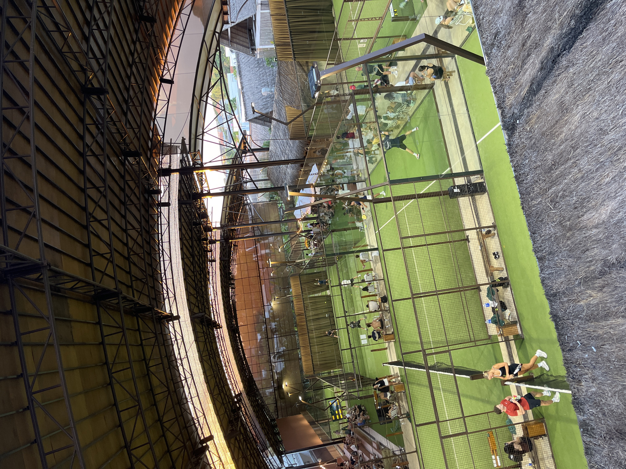 The image shows an overhead view of multiple padel tennis courts located in North Kuta, Indonesia. The courts are surrounded by glass walls and green turf, with players visible in various stages of a match. Some players are actively engaged in play while others are seated on benches. Spectators are seated near the courts, observing the matches. Overhead, a complex metal structure forms a partial roof, with the setting sun casting light onto its surface. Adjacent to the courts, there are structures resembling traditional Indonesian architecture with thatched roofs. A grassy area is visible in the foreground.