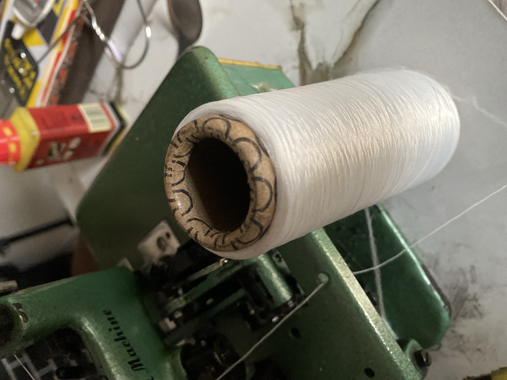 The media file shows a close-up, high-angle shot of a spool of white thread resting on a green industrial sewing machine. The spool is made of cardboard and is tightly wound with white thread. The sewing machine is green with some visible metal parts. The text "Machine" is faintly visible on the side of the sewing machine. In the background, out of focus, are other objects, including a red and yellow tin or container and some metallic items. The lighting suggests an indoor setting, possibly a workshop or a home craft area. The image does not contain any people or explicitly indicate the time of day or weather. The overall scene depicts a detail of a sewing setup, with a focus on the thread and the machine. The provided location context is Jauro Yinu, Nigeria, but this information is not visually discernible from the image itself.