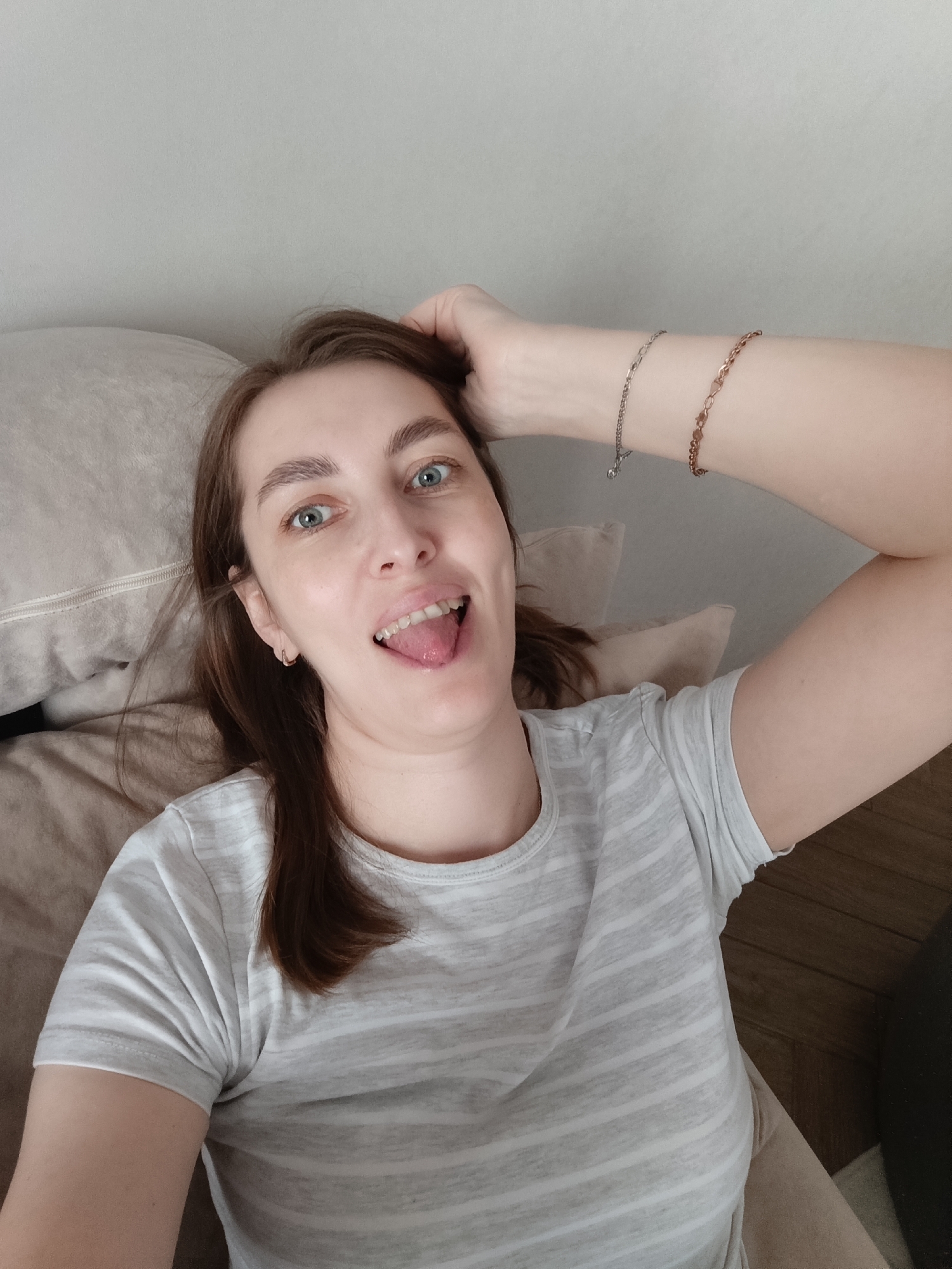 This is a self-portrait taken indoors, showing a young woman lying down in what appears to be a bedroom or living room in Chaikovsky, Russia. She is looking directly at the camera with a playful expression, her tongue slightly sticking out. Her right hand is raised, with fingers gently touching her hair near her forehead, revealing two chain bracelets on her wrist – one silver-toned and one rose gold-toned.
The woman has fair skin, long brown hair that falls around her shoulders, and light blue or green eyes. She is wearing a light grey t-shirt with horizontal white stripes. Her head rests on a beige or light brown pillow, which is part of a bed or couch. The background consists of a plain light grey wall. In the lower right corner, a dark brown wooden surface, possibly flooring or furniture, is visible. The lighting is soft and natural, suggesting it is daytime. The overall mood of the image is casual and relaxed. No text is visible in the image.