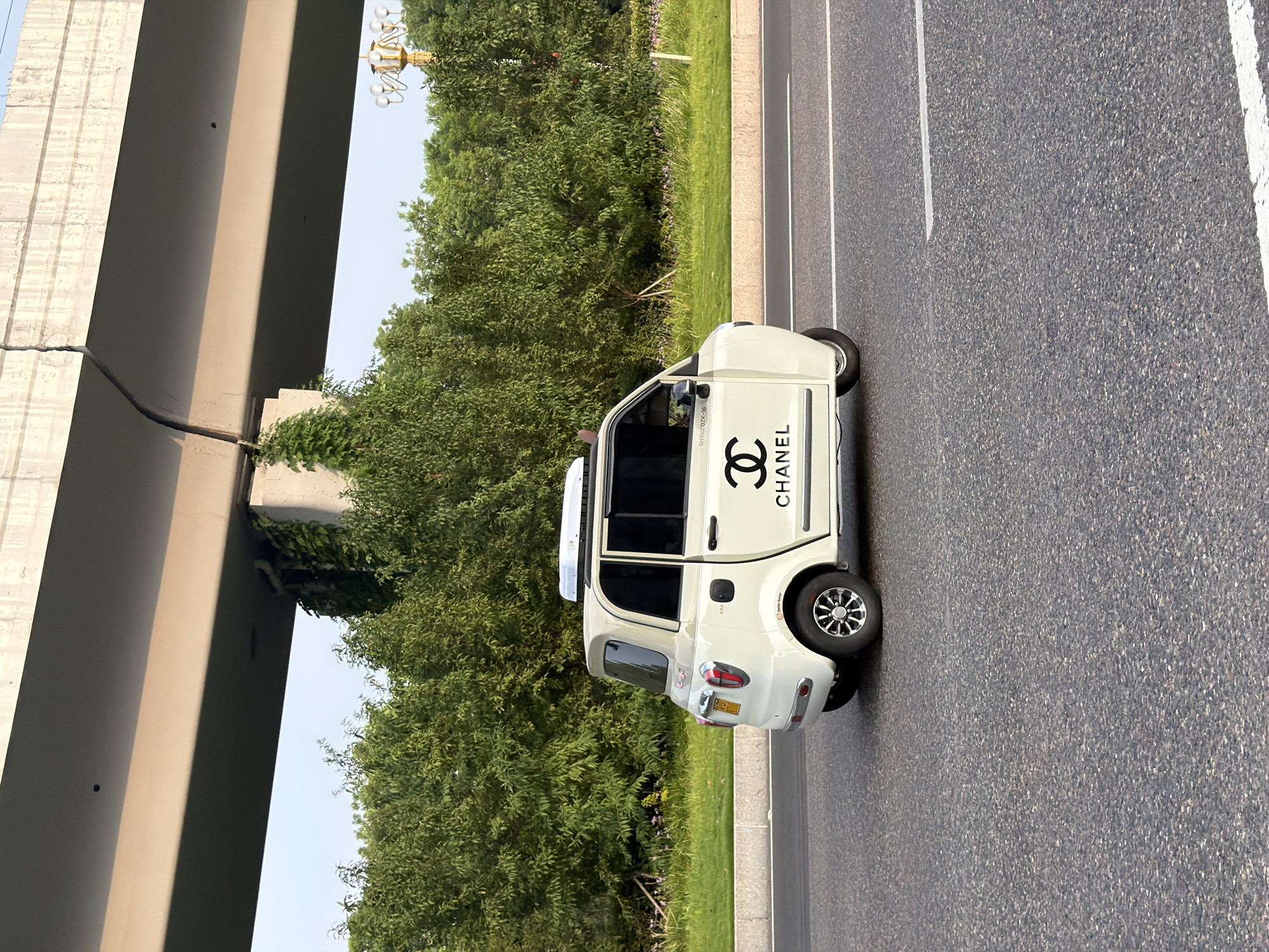An ivory-colored, three-wheeled electric vehicle, branded with the Chanel logo, drives along an asphalt road in Jiangjin District, Chongqing, China. The car, adorned with the iconic double 'C' logo and the word "CHANEL" in black lettering on its side, features black-tinted windows and a white roof-mounted cargo box. In the background, lush green trees line the roadside, and an elevated concrete overpass looms overhead. The sky is clear and bright, suggesting daytime. The scene appears to be a street view in a semi-urban or suburban area of Jiangjin.
