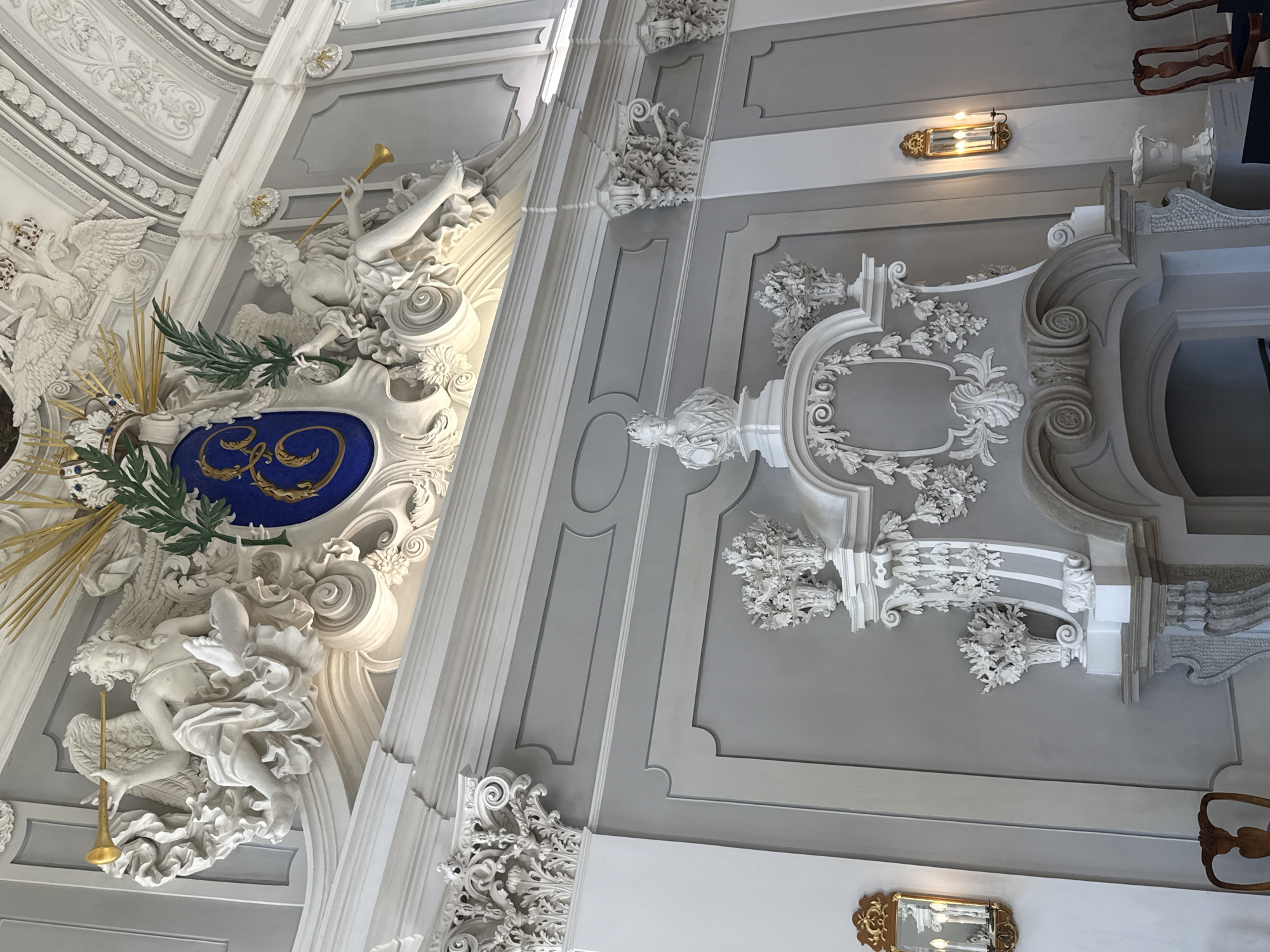 This media file captures a detailed view of ornate architectural detailing within what appears to be a grand interior, likely a church, palace, or similar historical building. The camera angle is looking upwards, emphasizing the ceiling and upper walls, which are richly decorated with stucco work.
The primary subjects are the elaborate sculptures and decorative elements. On the left side, there is a large stucco depiction of a winged figure, possibly an angel or cherub, blowing a golden trumpet. This figure is intertwined with a coat of arms or crest featuring a royal blue shield with golden embellishments, set against a background of golden rays and a green sprig. To the left of this, another large white stucco sculpture depicts what appears to be a swan or similar bird with a crown on its head.
The right side of the image showcases a different section of the interior, characterized by more of the white stucco ornamentation against a light gray painted wall. These decorations include intricate floral motifs, scrollwork, and framing elements. A notable object in this section is a small, rectangular wall sconce with a golden, ornate frame holding three light bulbs, casting a warm glow. Further down, a small, gilded mirror with intricate detailing is visible on a white wall. The overall impression is one of opulence and historical grandeur.
The scene suggests a static, internal environment, with no indication of a specific time of day or weather conditions. The focus is entirely on the artistic and architectural features. There is no visible text within the image. The overall mood is one of awe and admiration for the craftsmanship.