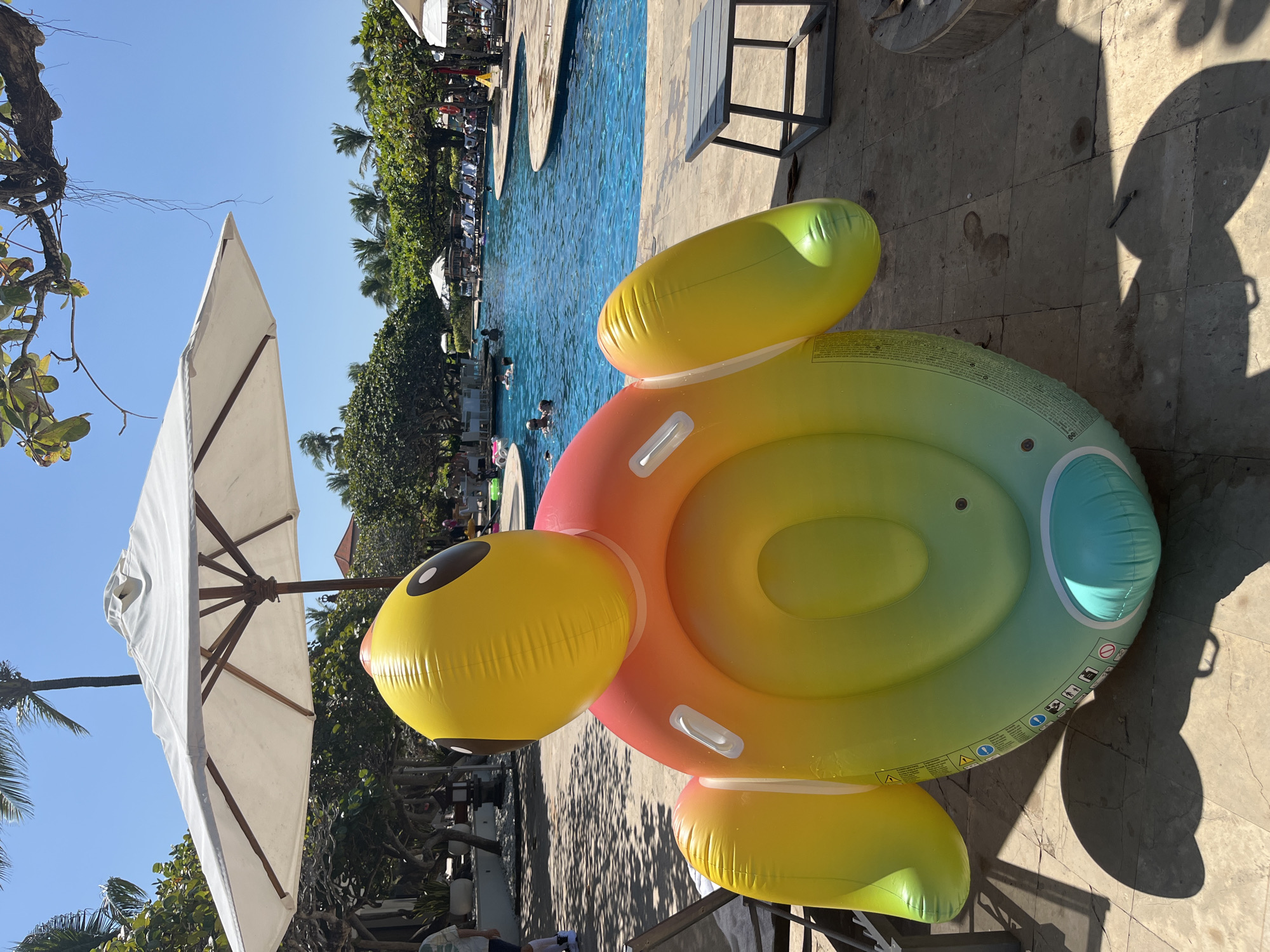 The image depicts an inflatable turtle pool float positioned on a tiled patio area next to a swimming pool. The float is brightly colored, transitioning from yellow at the top to orange, pink, and green towards the bottom. It has a large round opening in the center and two arm-like extensions on either side of its head. Behind the float, a large blue swimming pool is visible, with several people swimming and relaxing in and around it. Palm trees and lush green foliage surround the pool area, suggesting a tropical or subtropical environment. In the background, there are lounge chairs and umbrellas, typical of a resort or hotel setting. The sky is clear and blue, indicating a sunny day. The location is identified as Kuta Selatan, Indonesia, which is known for its beach resorts and vacation destinations. The scene conveys a sense of leisure and relaxation.