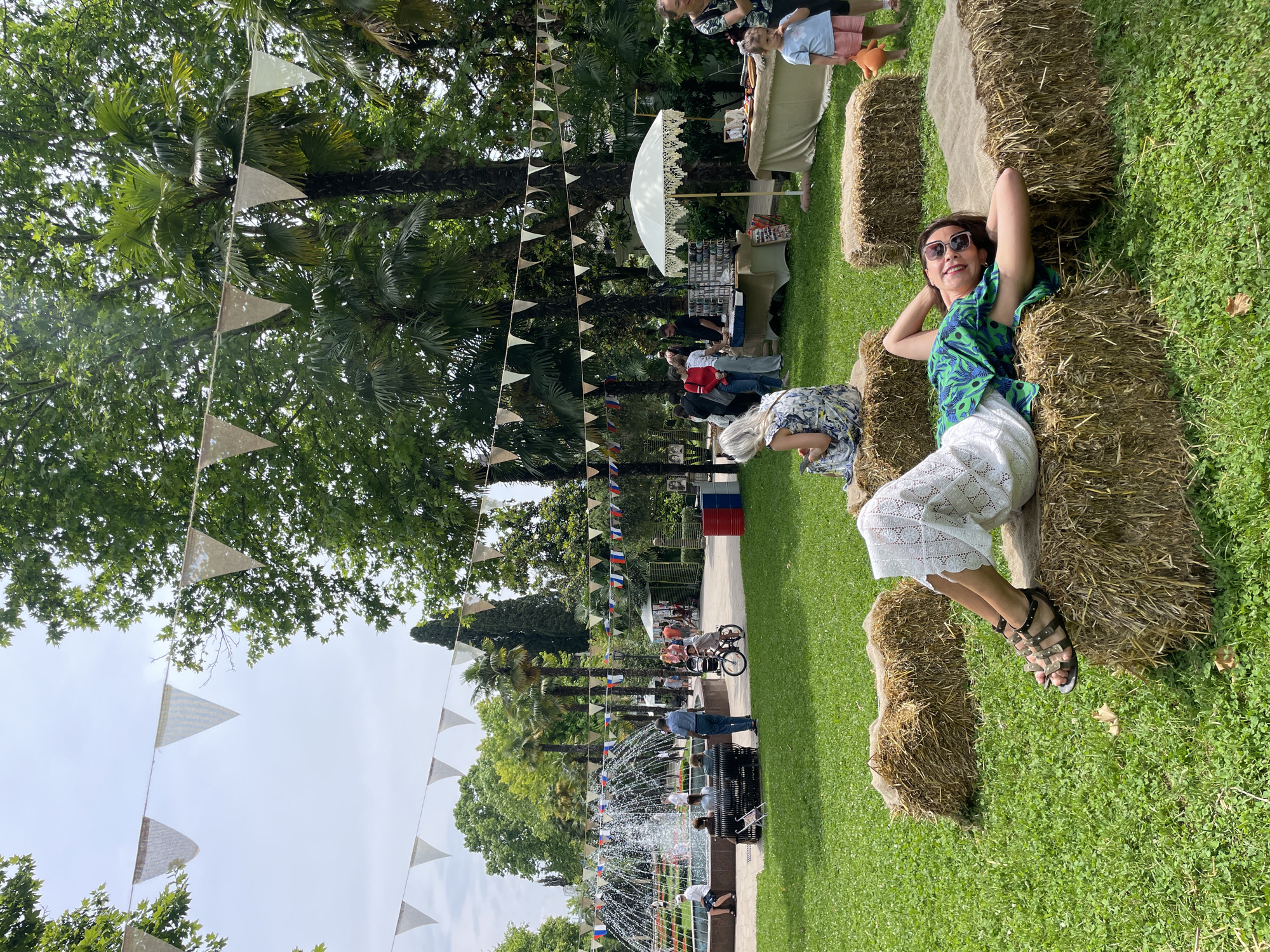 The image depicts an outdoor event, possibly a fair or market, set in a park in Sochi, Russia. In the foreground, a woman with dark hair and sunglasses is lying down on a hay bale, resting her head on her arms. She is wearing a green top with a blue floral print and a white, lacy skirt. She is smiling and looking towards the camera. Several other hay bales are scattered around the grassy area.
In the background, a fountain is visible with water spraying upwards. People are gathered around the fountain and walking along a paved pathway. Several market stalls with white canopies are set up, displaying various items. Bunting is strung between trees, decorating the scene. The environment is lush with green grass, trees, and palm trees, suggesting a warm climate. The overall atmosphere appears relaxed and festive.